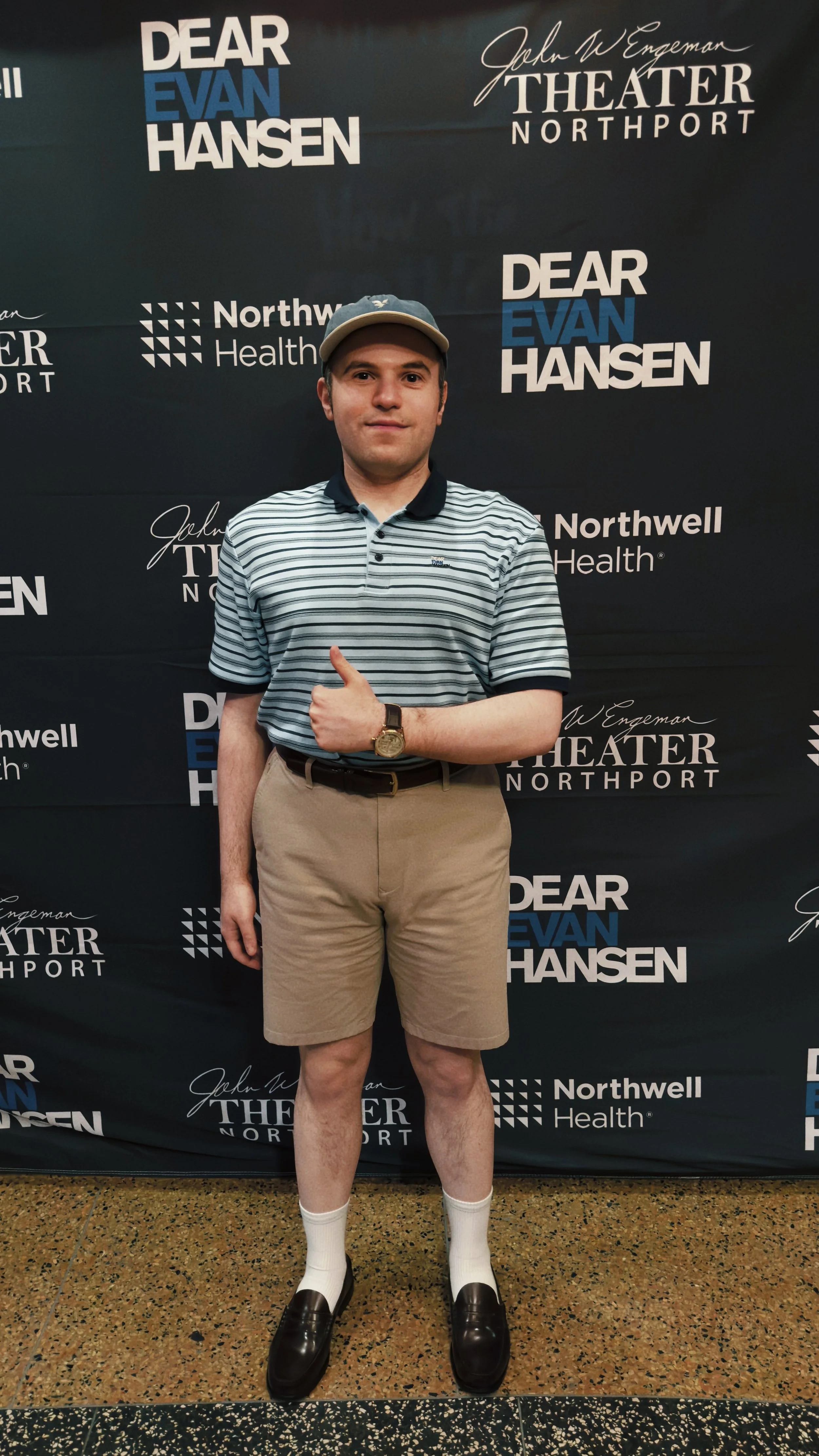 Smiling man in a striped polo, khaki shorts, and cap gives a thumbs-up at a Dear Evan Hansen theater event backdrop.