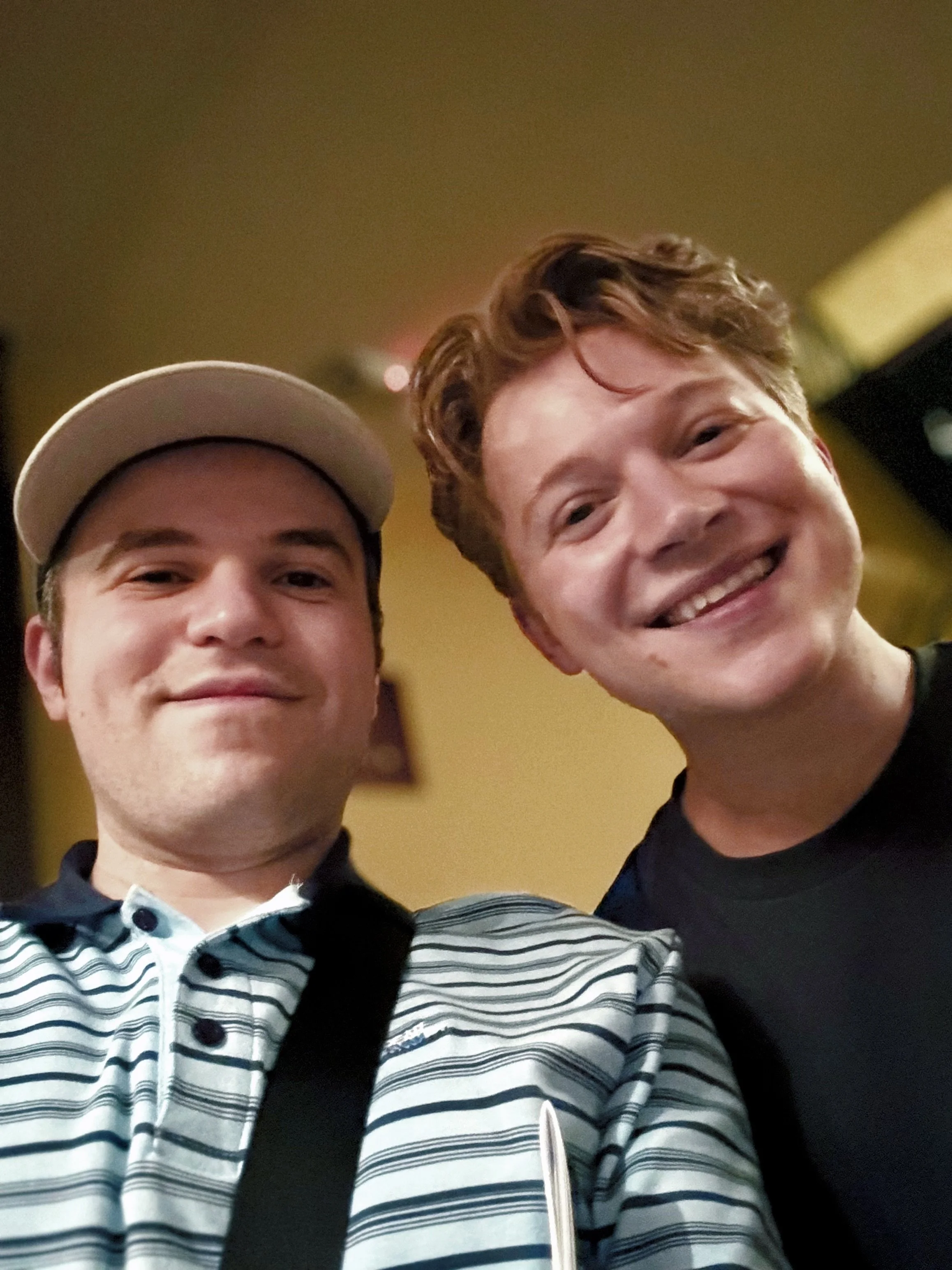 Two smiling friends take a close-up indoor selfie; one wears a cap and striped shirt, while the other wears black, creating a warm, casual mood.