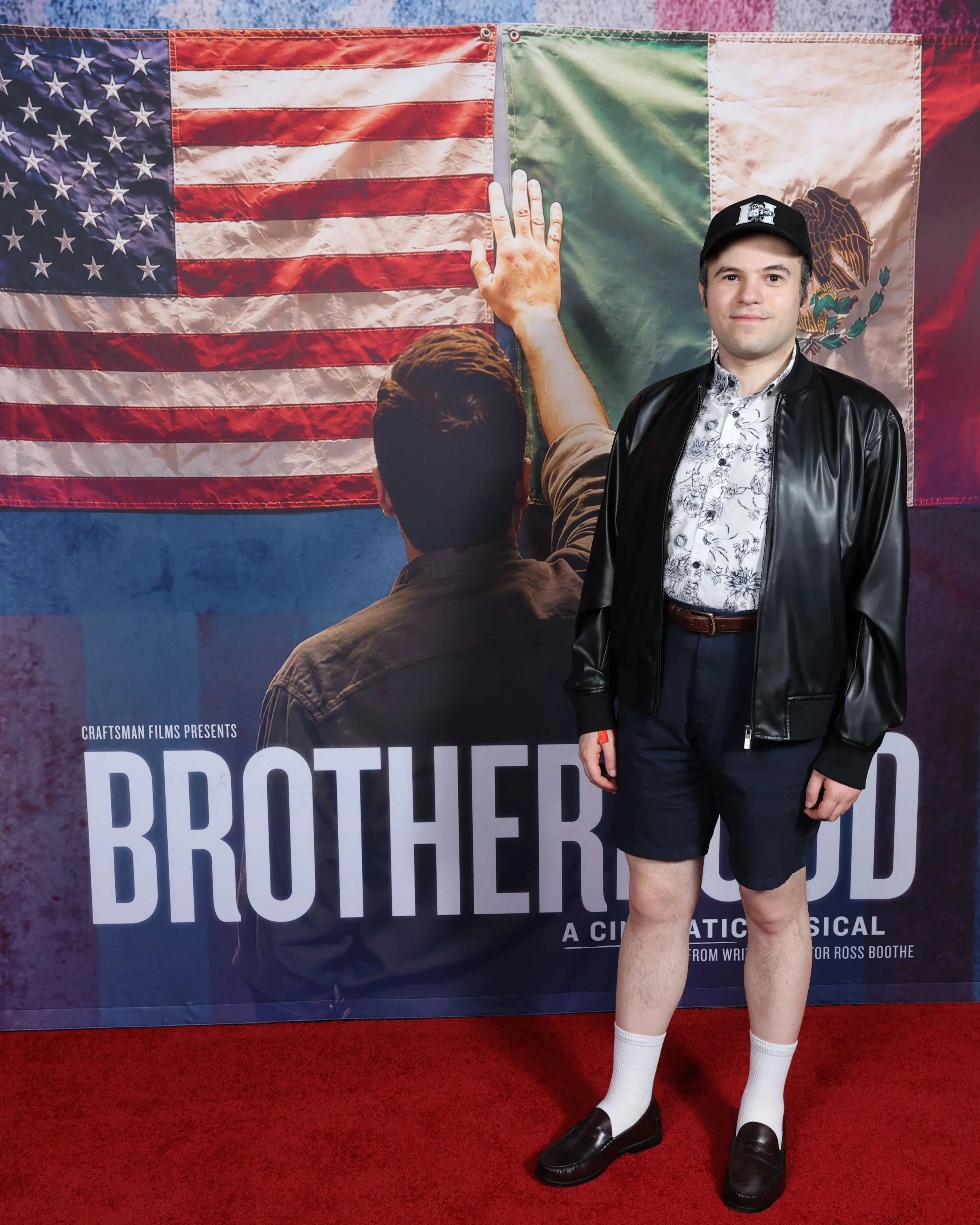 Man in a black leather jacket, patterned shirt, shorts, and cap poses on a red carpet before a “Brotherhood” musical poster featuring American and Mexican flags.