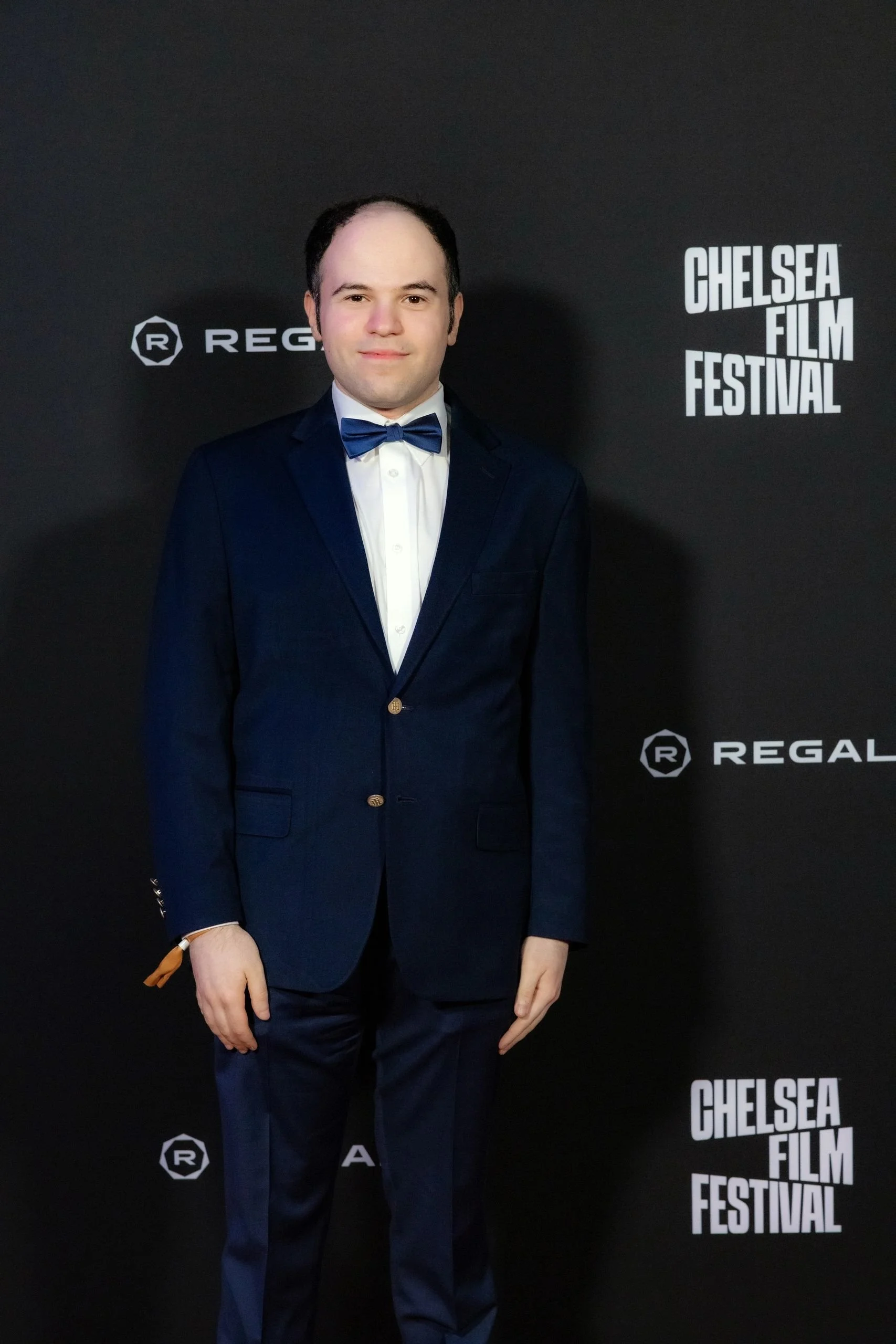 Man in navy suit, white shirt, and blue bow tie standing on a black carpet at the Chelsea Film Festival.