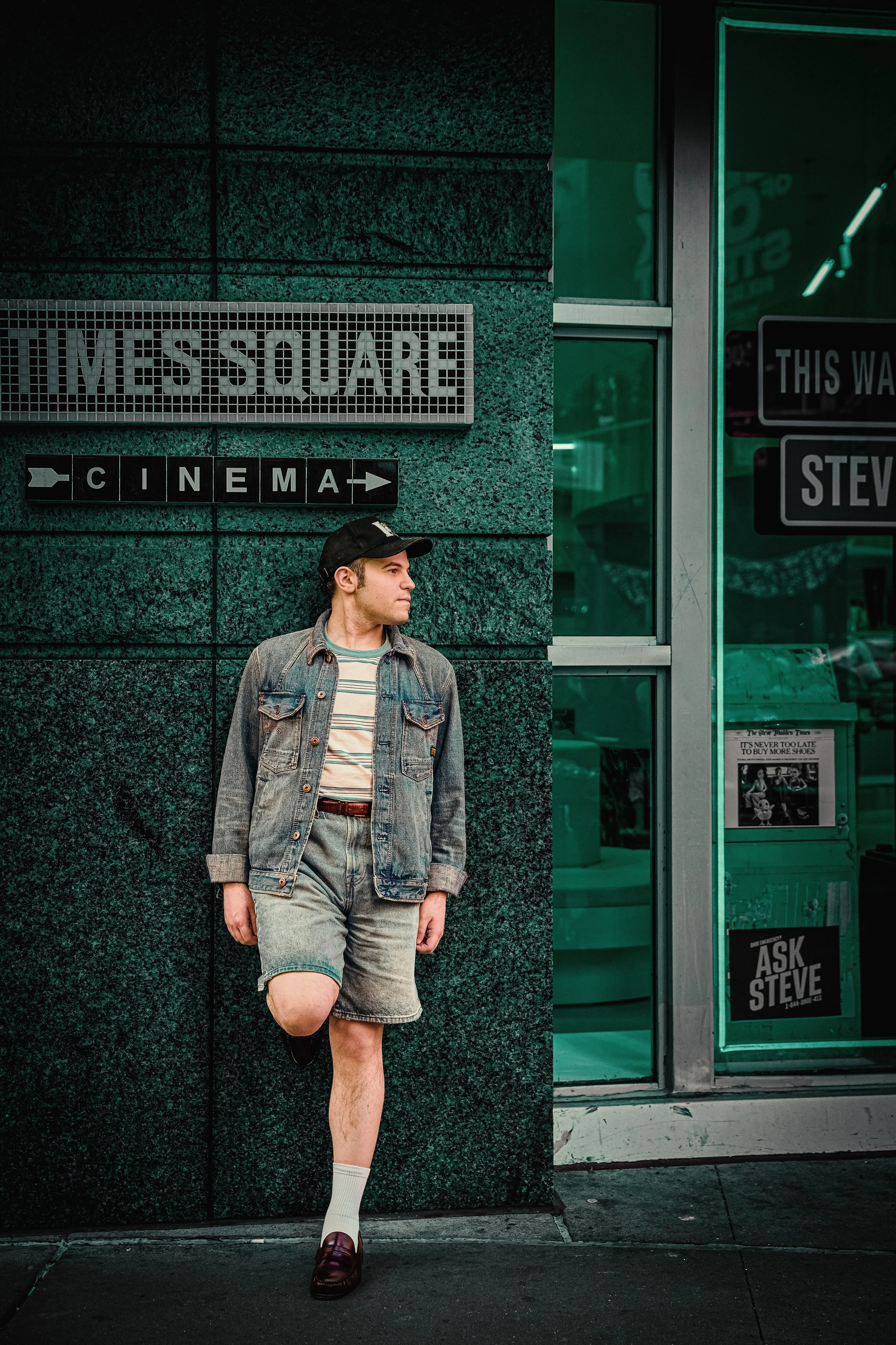 Young man in a denim jacket, cap, and shorts leans against a teal theater wall beneath “Times Square Cinema” signage in a moody urban portrait.