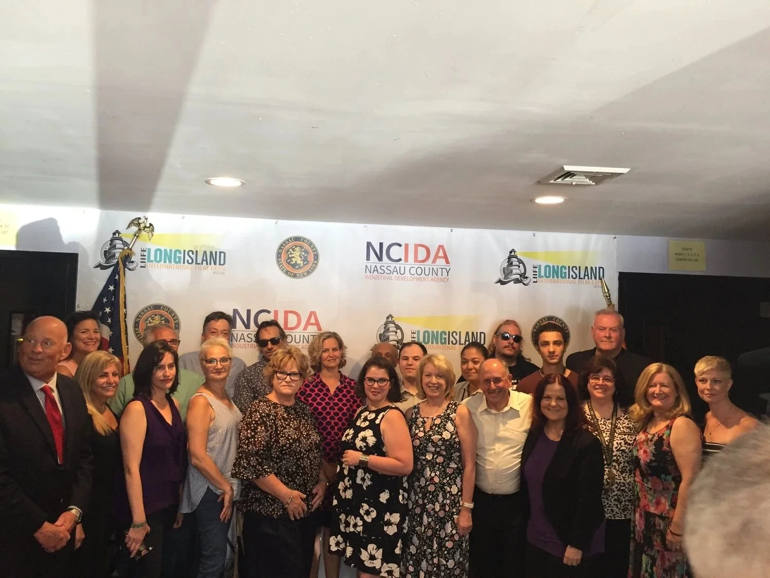 Group of people posing together at an event with a backdrop featuring logos and banners related to Long Island and Nassau County.