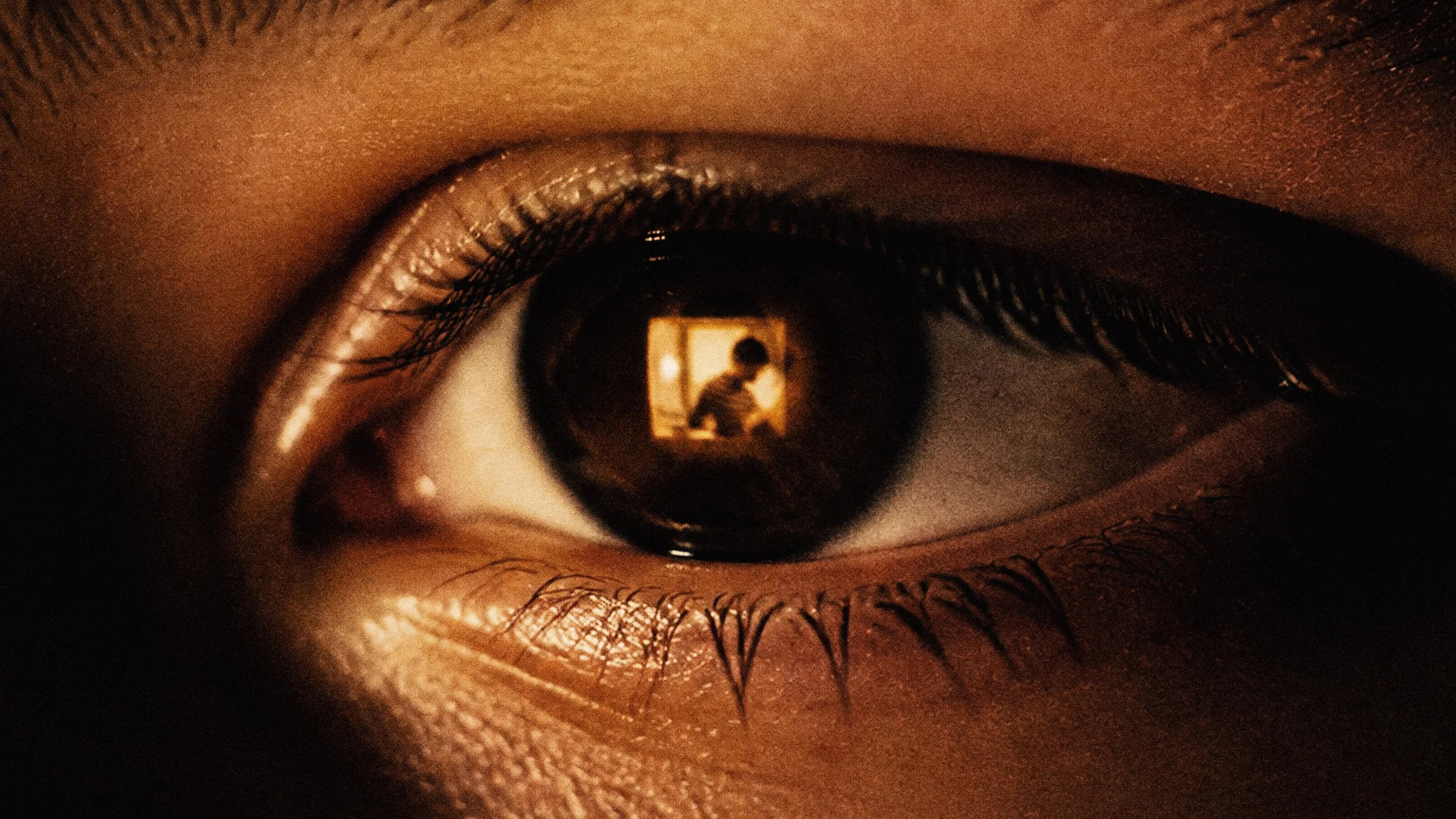 Extreme close-up of a dark brown eye with long lashes, warm amber lighting, and a small silhouetted figure reflected in the pupil, creating a moody tone.