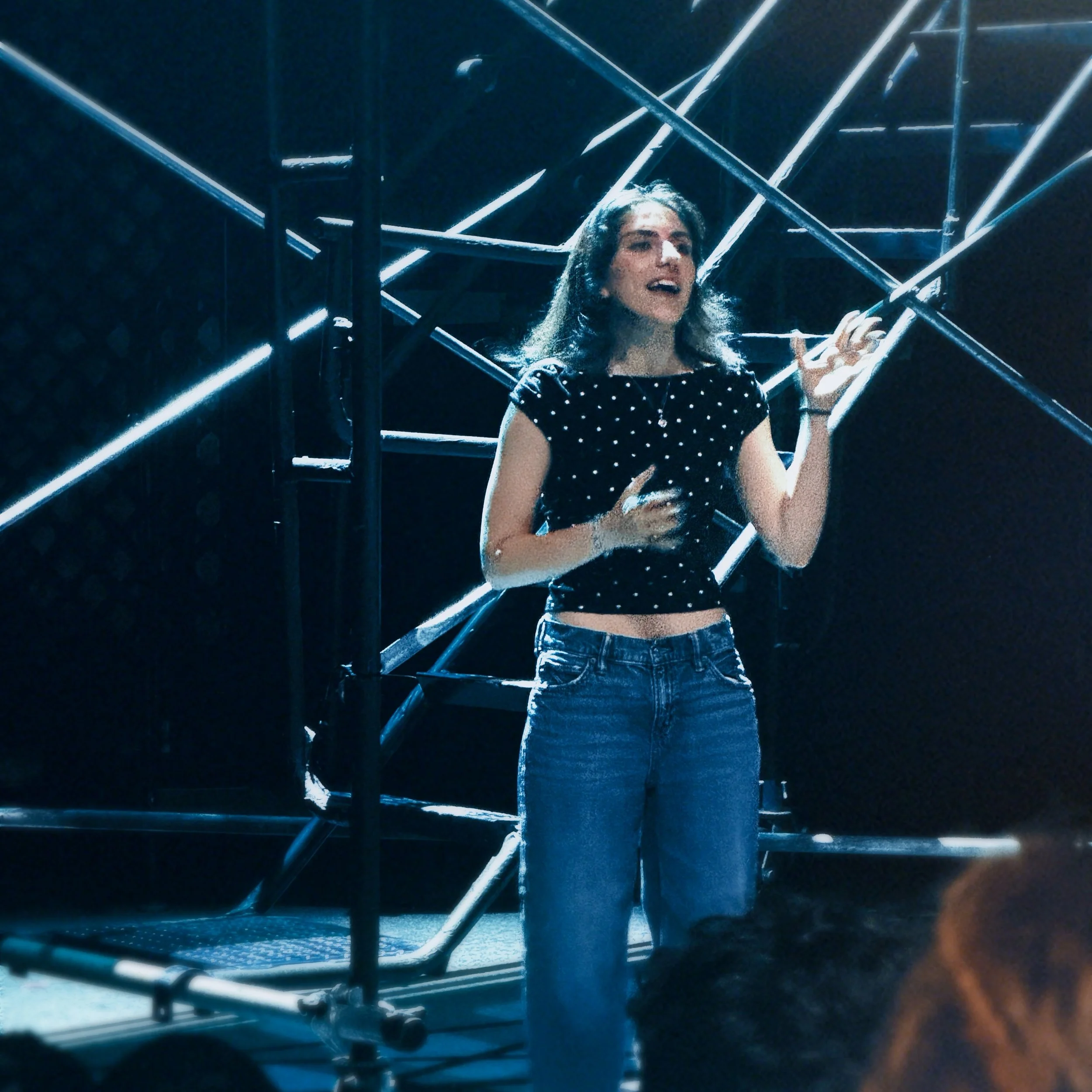 A young woman in a polka-dot crop top and jeans speaks animatedly on a dimly lit stage with a metal scaffolding background.