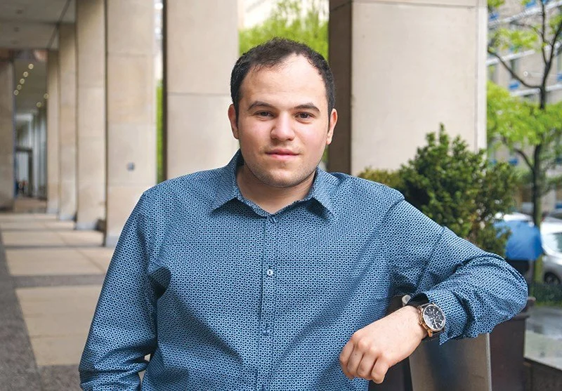A young man with short dark hair wearing a blue patterned shirt, standing outdoors with a railing, trees, and a city street in the background.