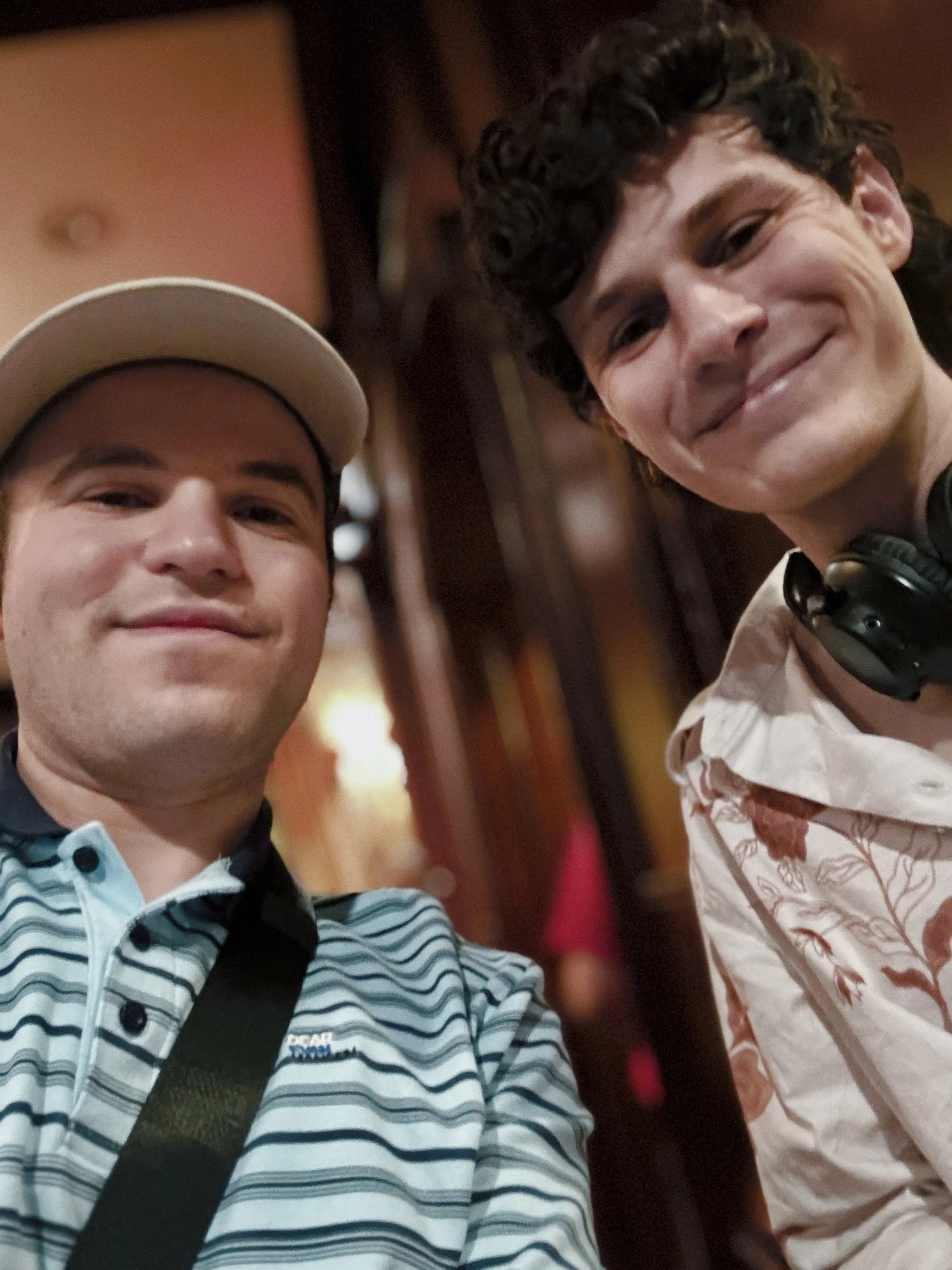 Two smiling young men take a casual selfie indoors; one wears a cap and striped shirt, while the other has headphones around his neck.