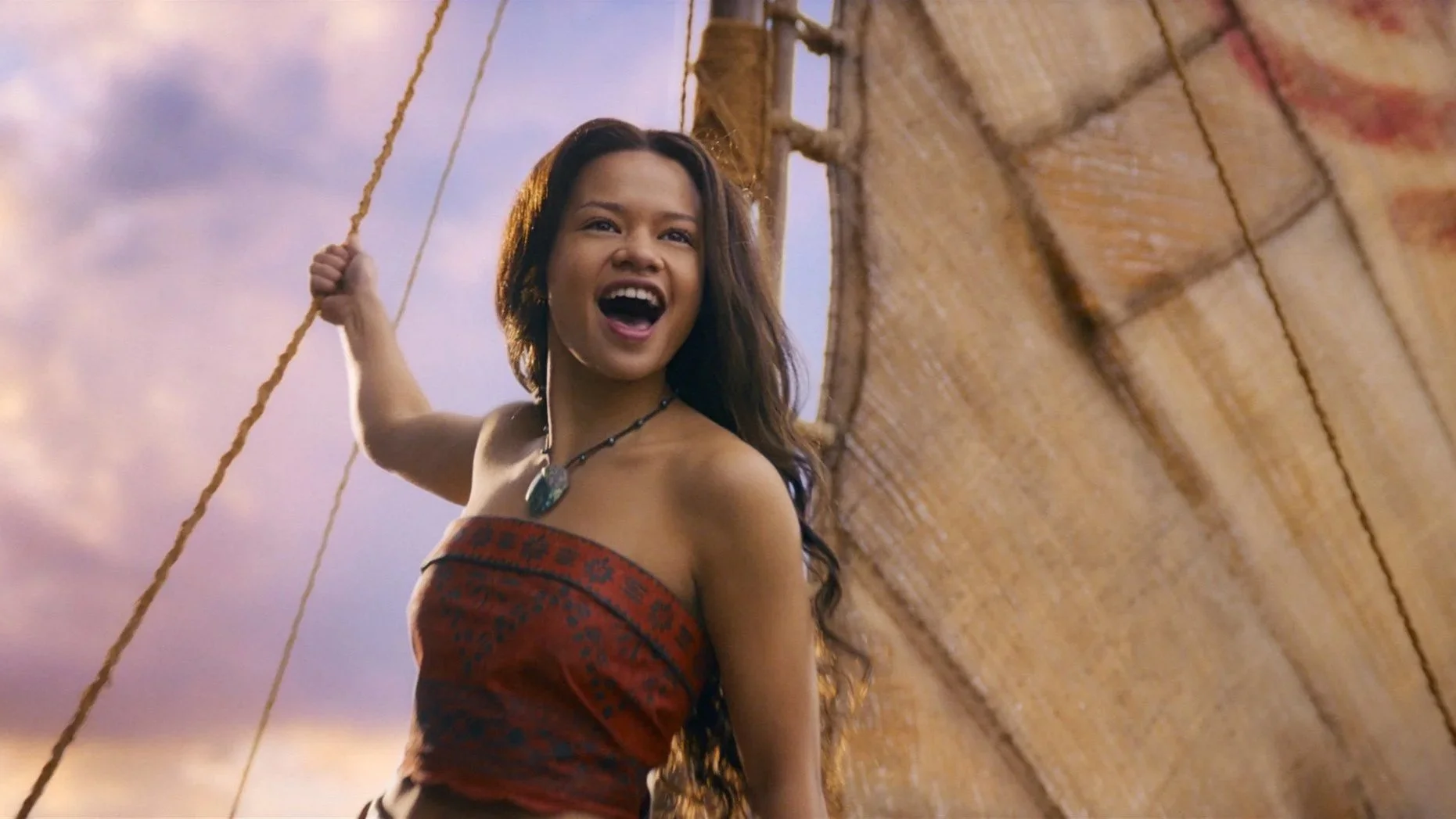 A woman smiling in a red dress outdoors, with a clear sky and fluffy clouds in the background. She is near water, possibly by a sailboat.