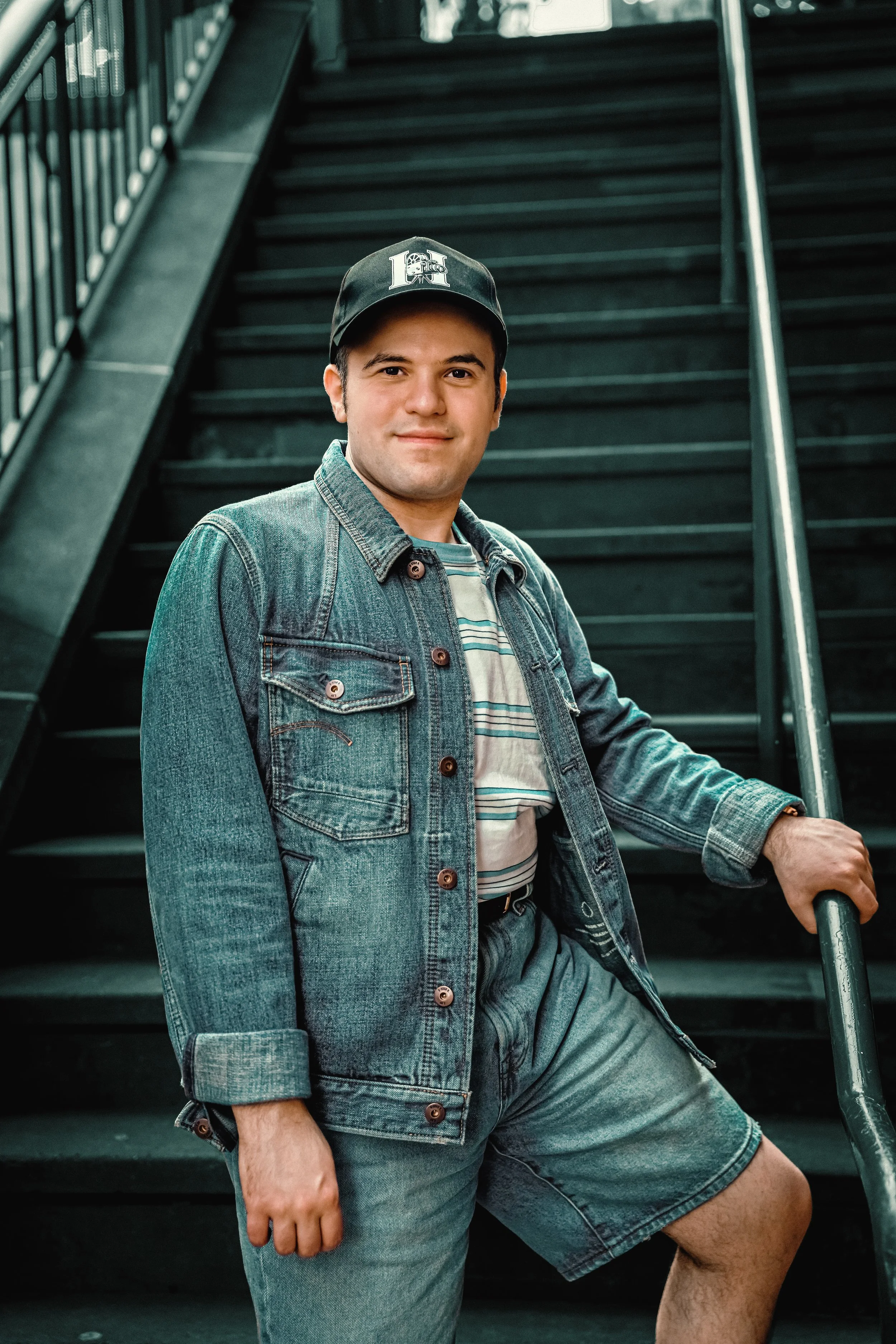 Smiling man in a denim jacket, shorts, and baseball cap poses on a dark stairway, one hand on the railing, conveying a relaxed, confident mood.