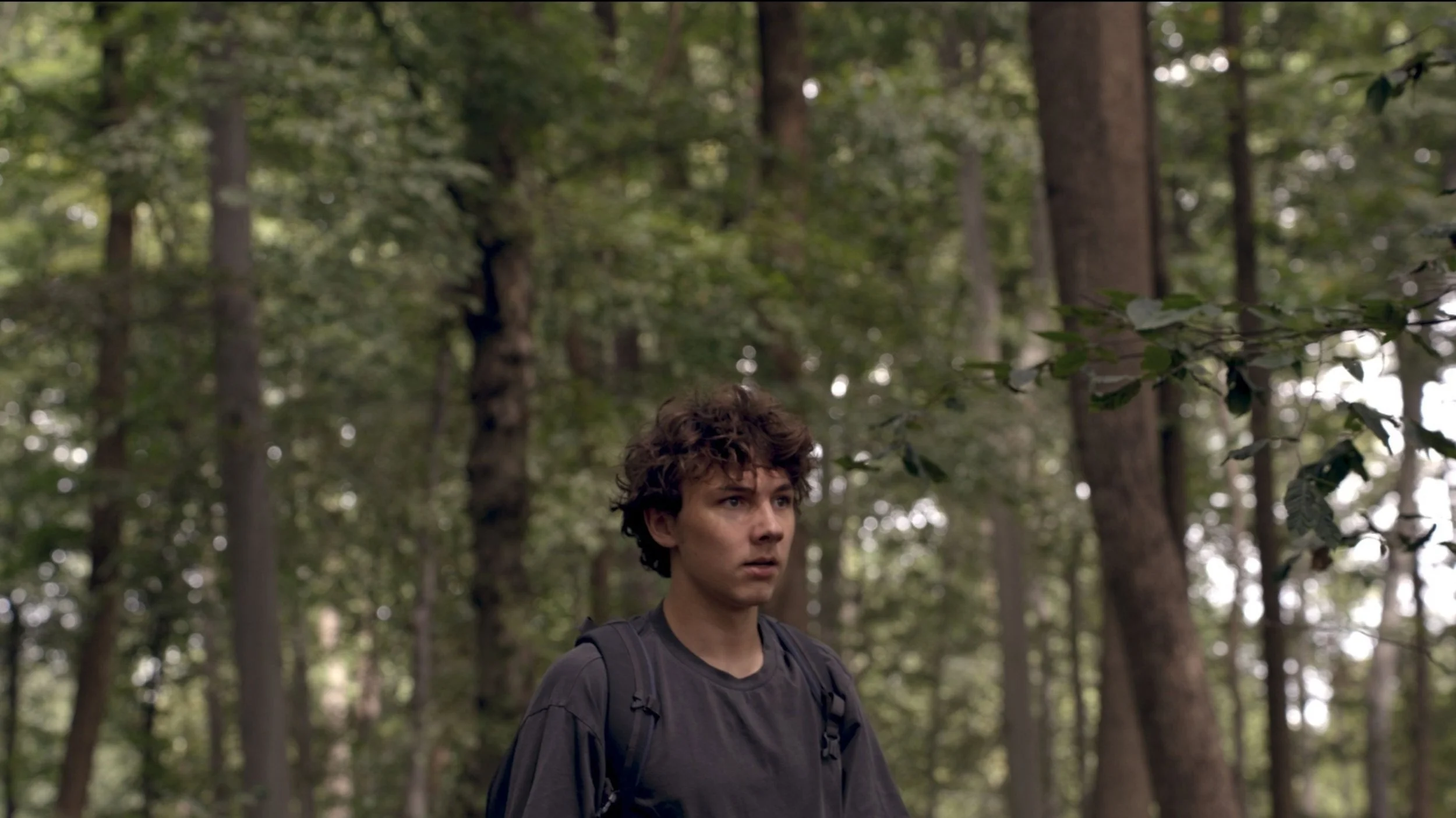 Young man with backpack walks through a dense woodland, his serious expression creating a quiet, contemplative cinematic mood.