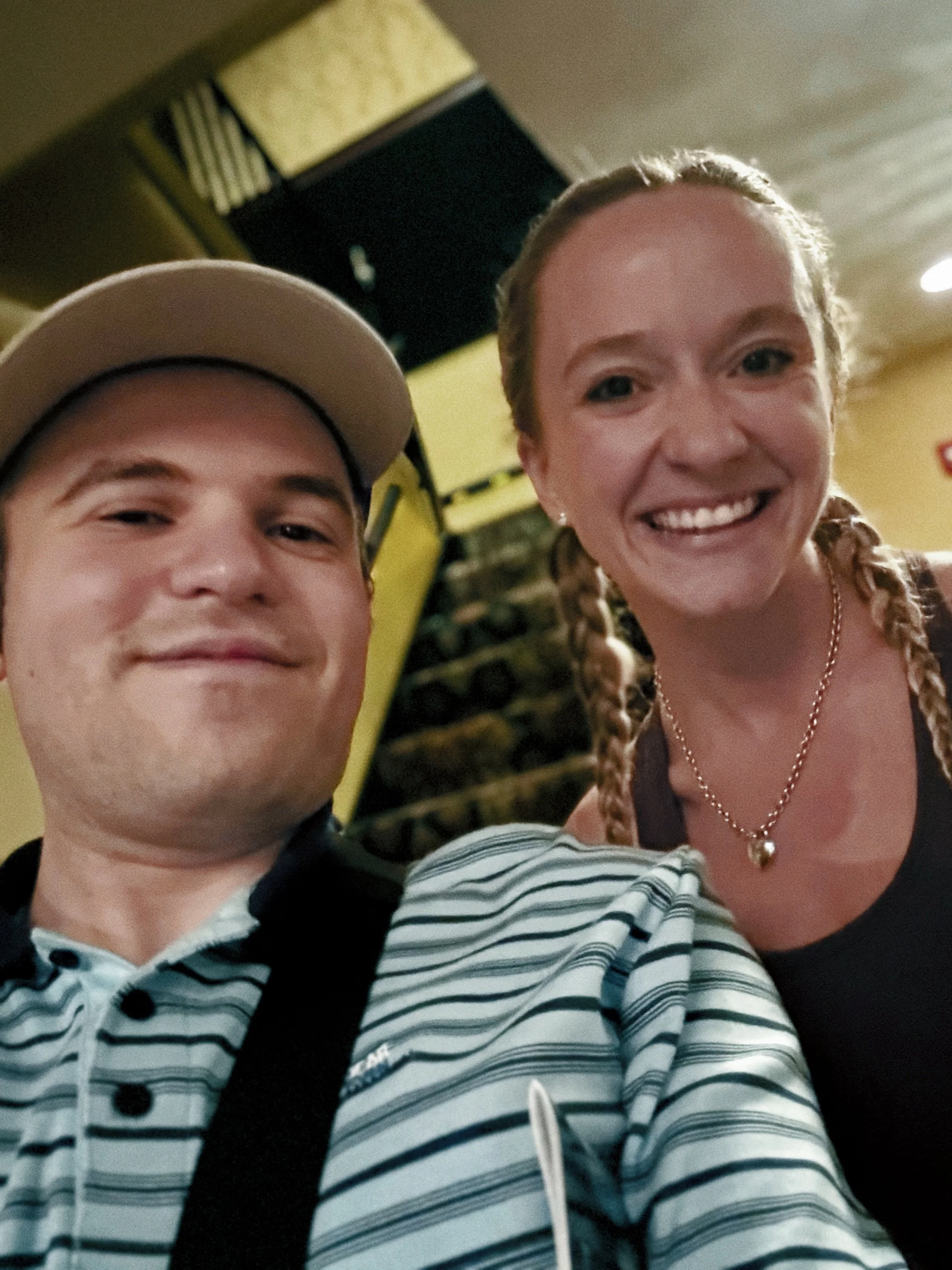 Smiling man in a cap and striped shirt takes a selfie with a smiling woman in braids indoors, creating a warm, friendly atmosphere.