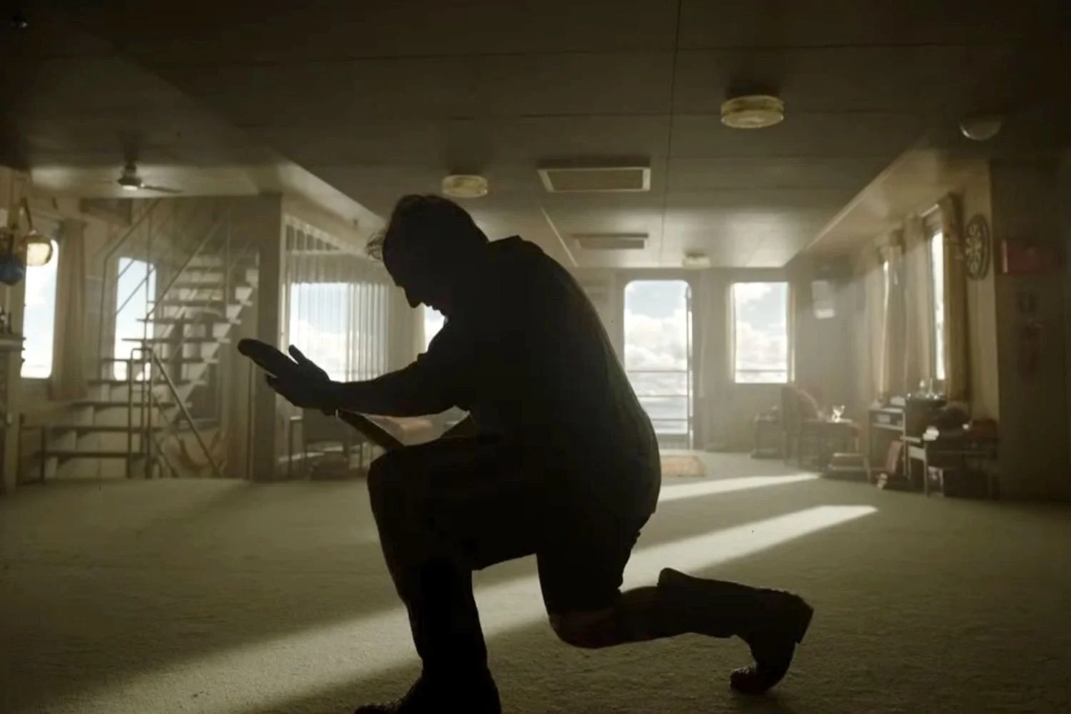 A man kneeling in a dimly lit room with walls, a ceiling, and a floor visible.