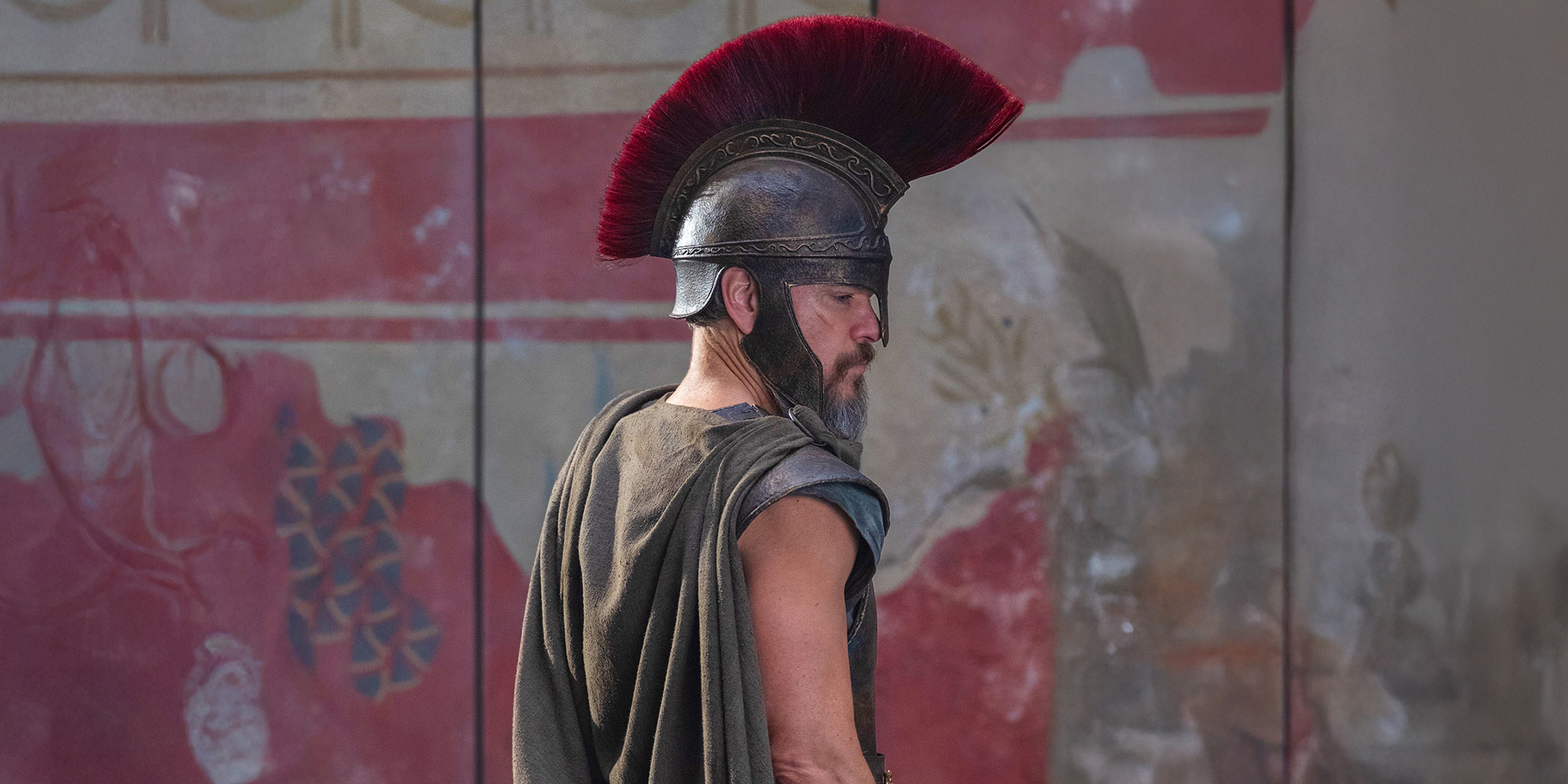 A man wearing ancient Greek or Roman armor and a helmet with a large red plume stands in front of a painted wall.