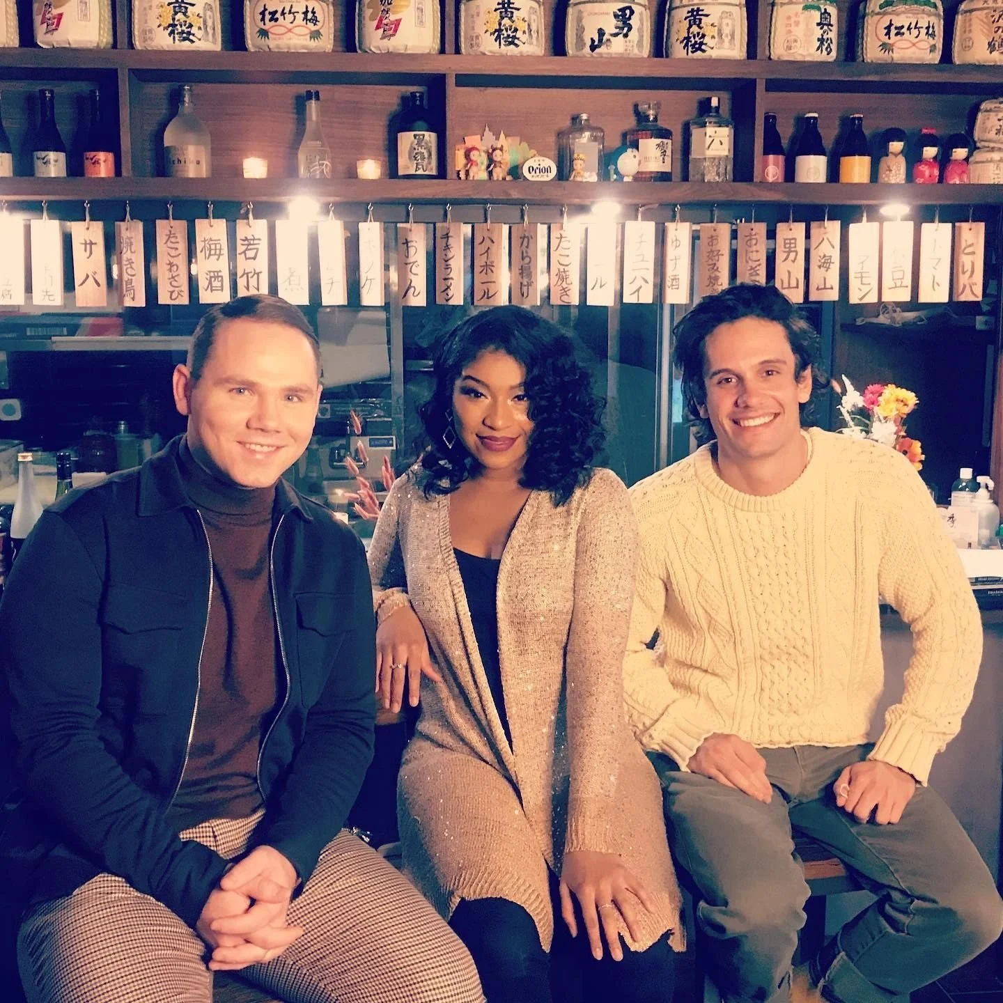 Three people sitting in front of a Japanese restaurant bar with bottles of alcohol and Japanese signs on the wall behind them.