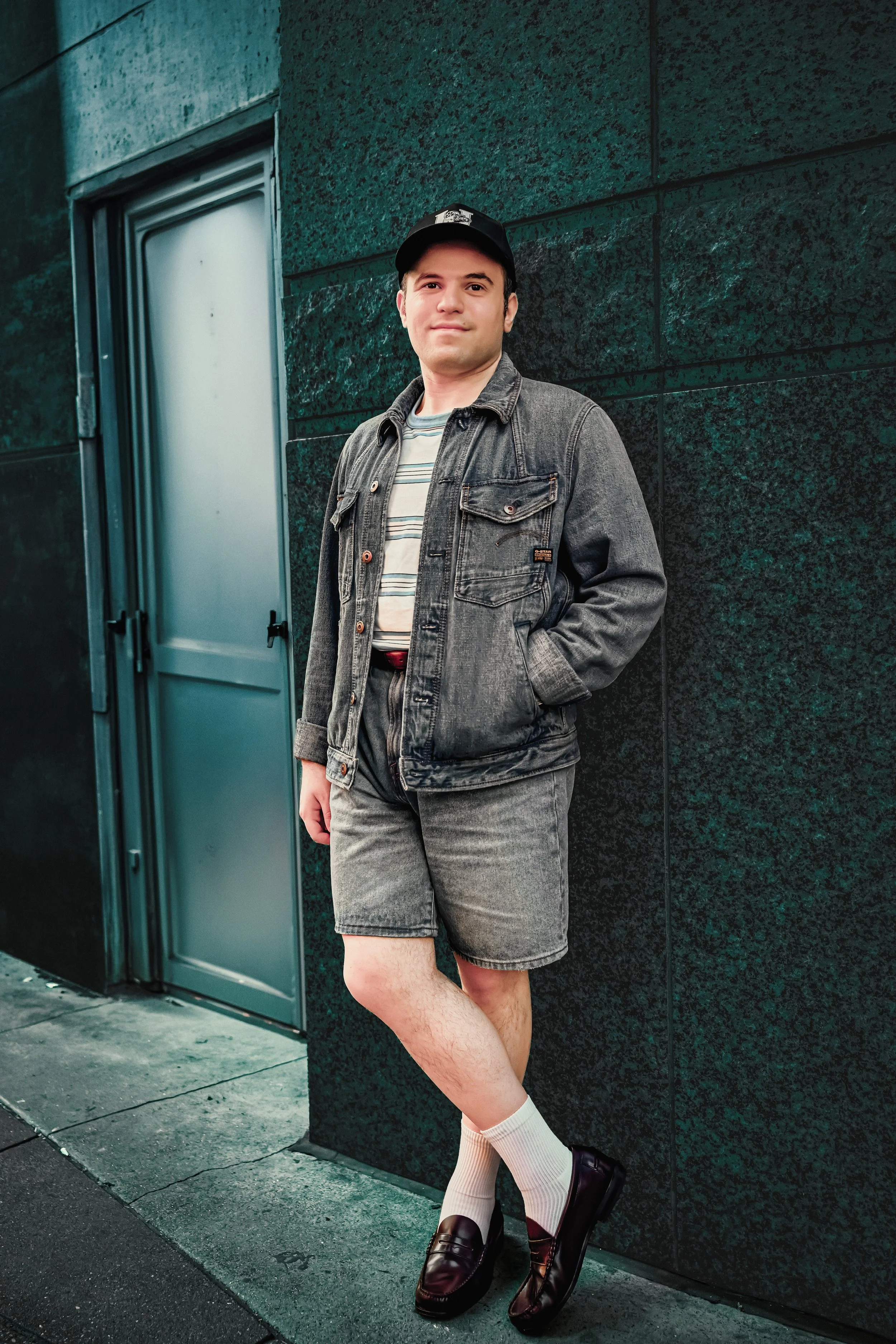 A relaxed man in a cap, denim jacket, striped shirt, and gray shorts leans against a dark tiled wall beside a teal door, smiling.