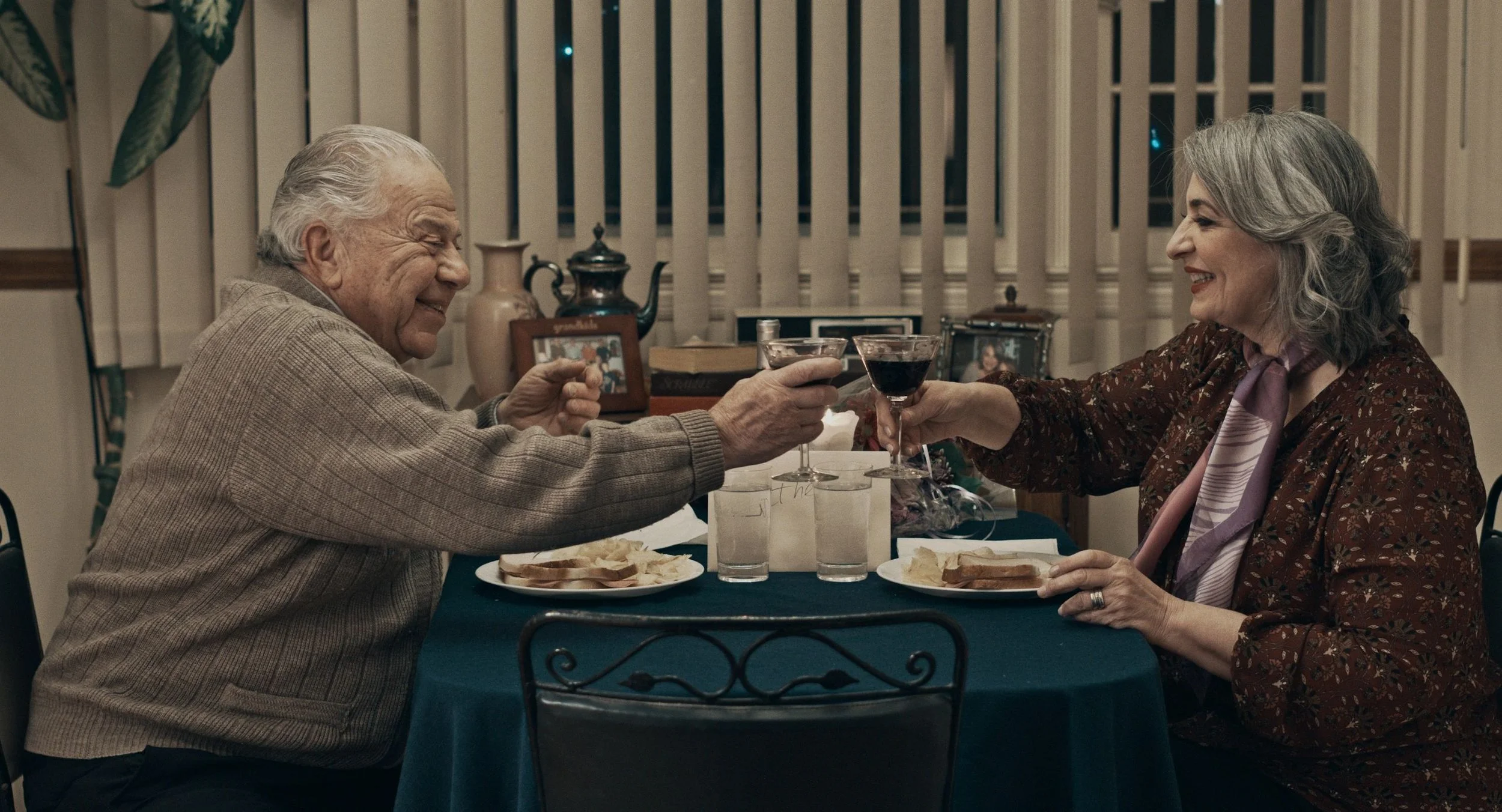 An elderly man and woman enjoying a toast at a dinner table. They are smiling, with the man on the left and the woman on the right, holding glasses of red wine. The table is set with plates of food, glasses of water, and decorative items in the background.