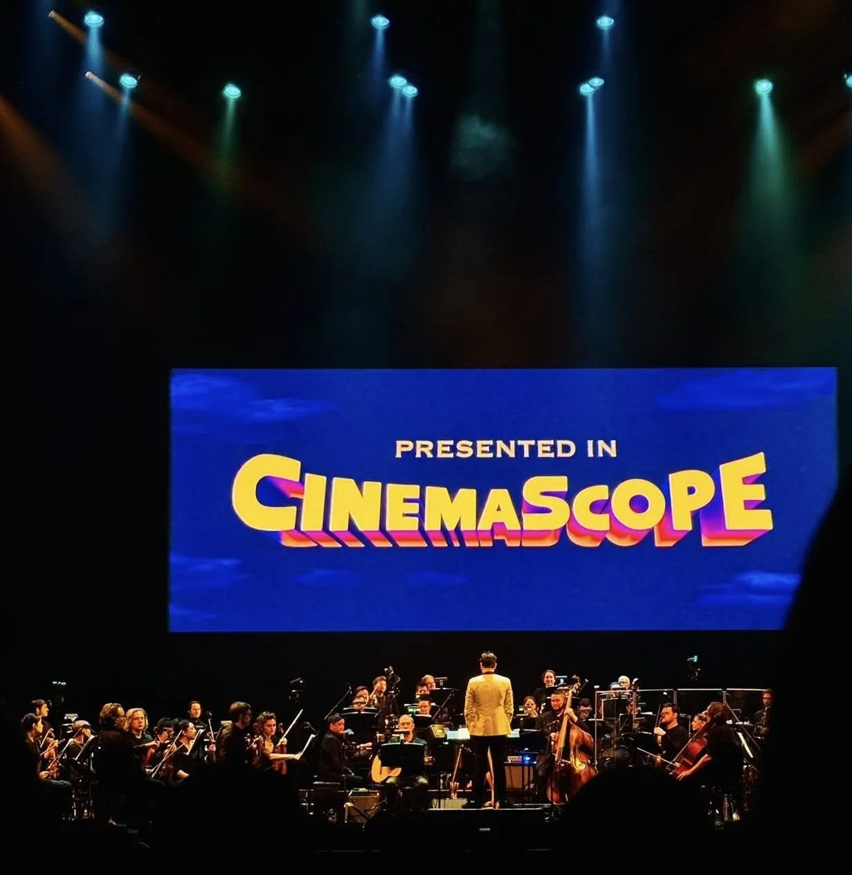An orchestra performs on stage under dramatic lighting, with a large screen above displaying the words "Presented in Cinemascope." The atmosphere is vibrant and grand.