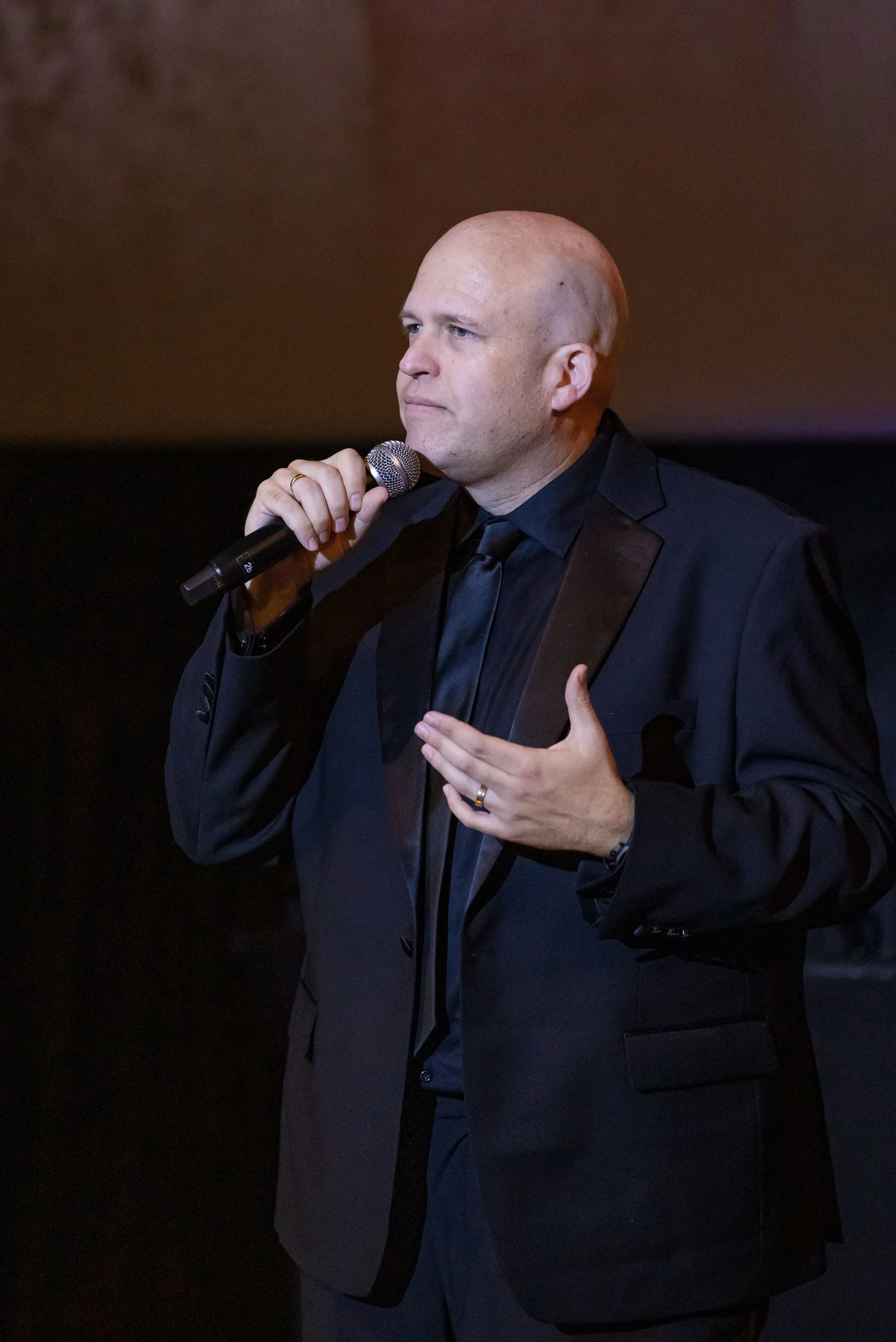 Bald man in a dark suit speaks into a handheld microphone, gesturing with his free hand against a dim stage backdrop; the mood is serious and focused.