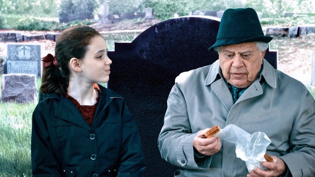 A young girl and an older man standing in a cemetery, with the man looking at a sandwich. The girl is looking at him, and the background shows headstones and grass.