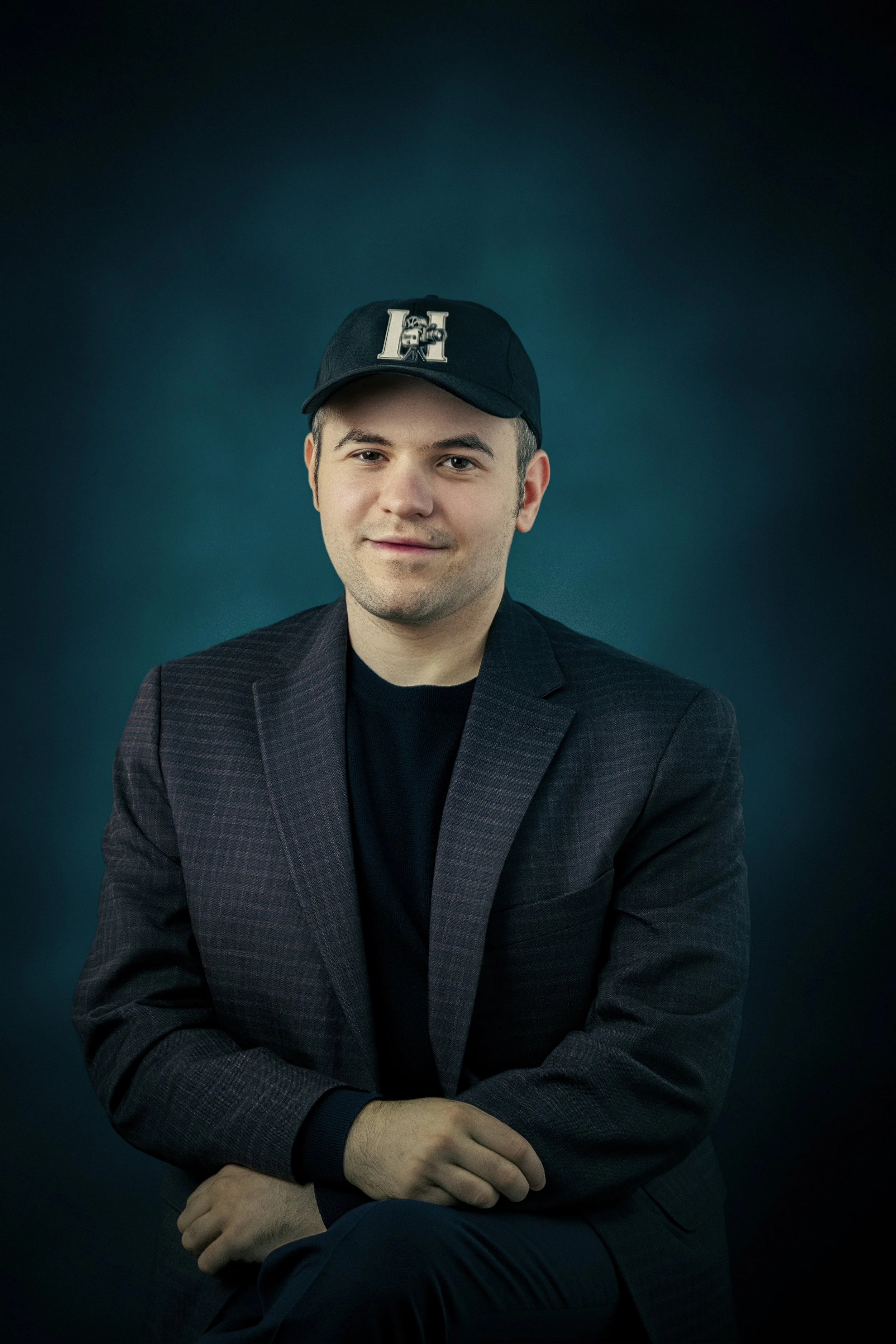 A young man with light skin wearing a dark blazer, black shirt, and a black baseball cap with a large 'H' on it. He is sitting with his arms crossed against a dark teal background, smiling slightly.
