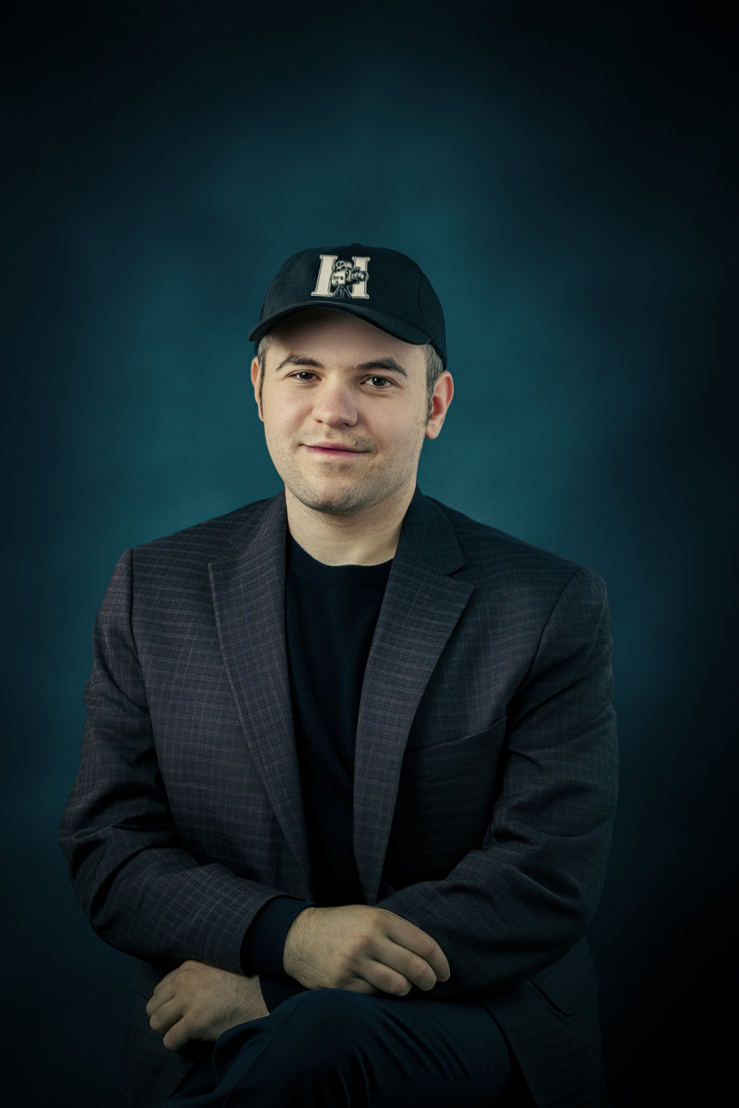 A young man with light skin wearing a dark blazer, black shirt, and a black baseball cap with a large 'H' on it. He is sitting with his arms crossed against a dark teal background, smiling slightly.