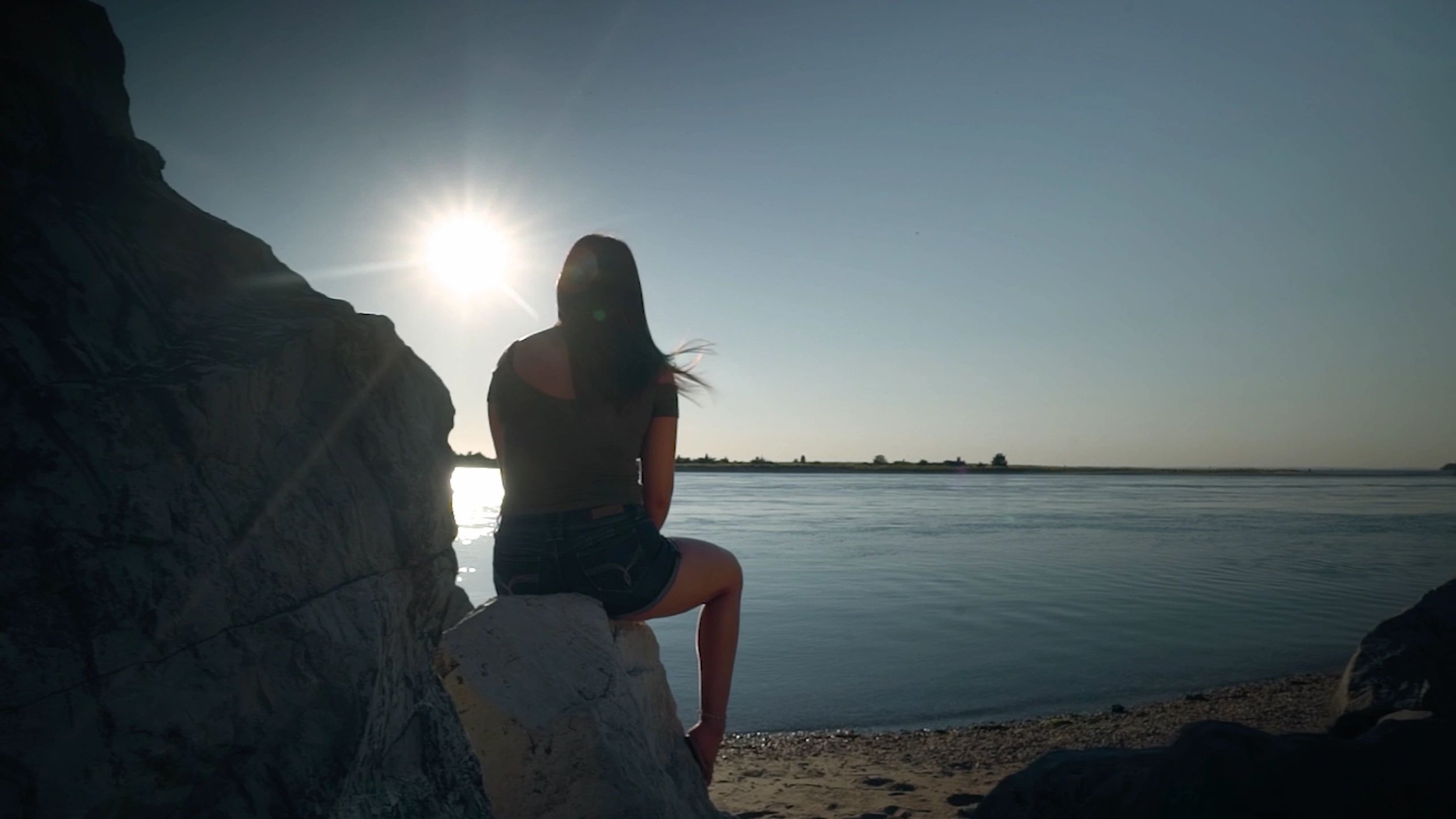 A woman sitting on a rock by the water during sunset, facing away from the camera. The sun is low in the sky, casting a glow on the scene, with water and distant land in the background.
