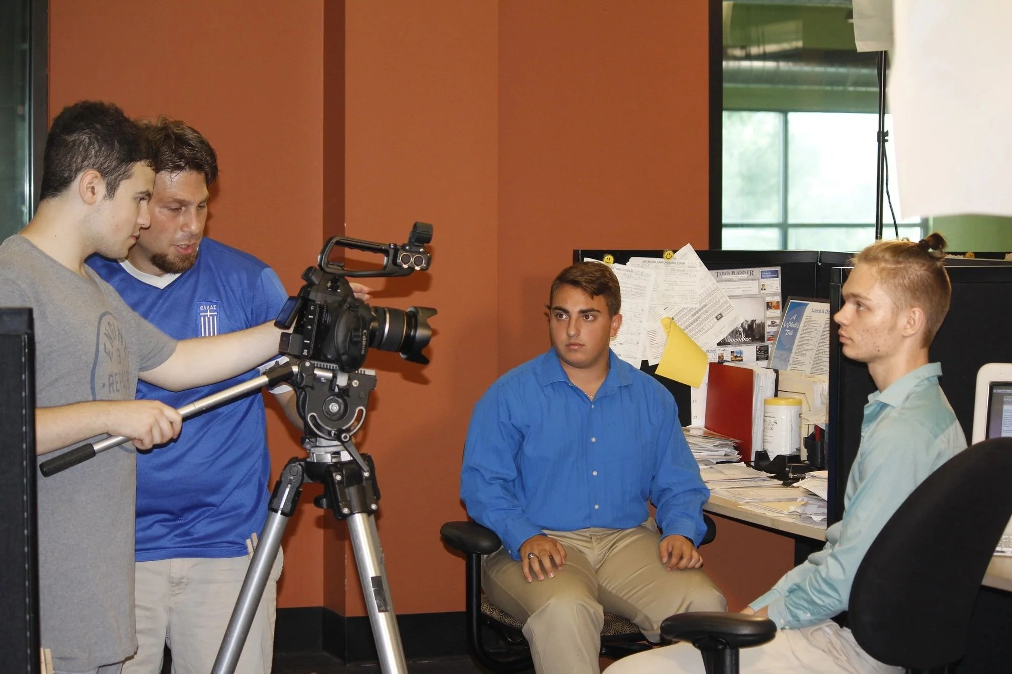 Two men are filming a scene with a camera in an office setting while two others are sitting and acting as if in an interview or discussion, with desks, papers, and a computer in the background.