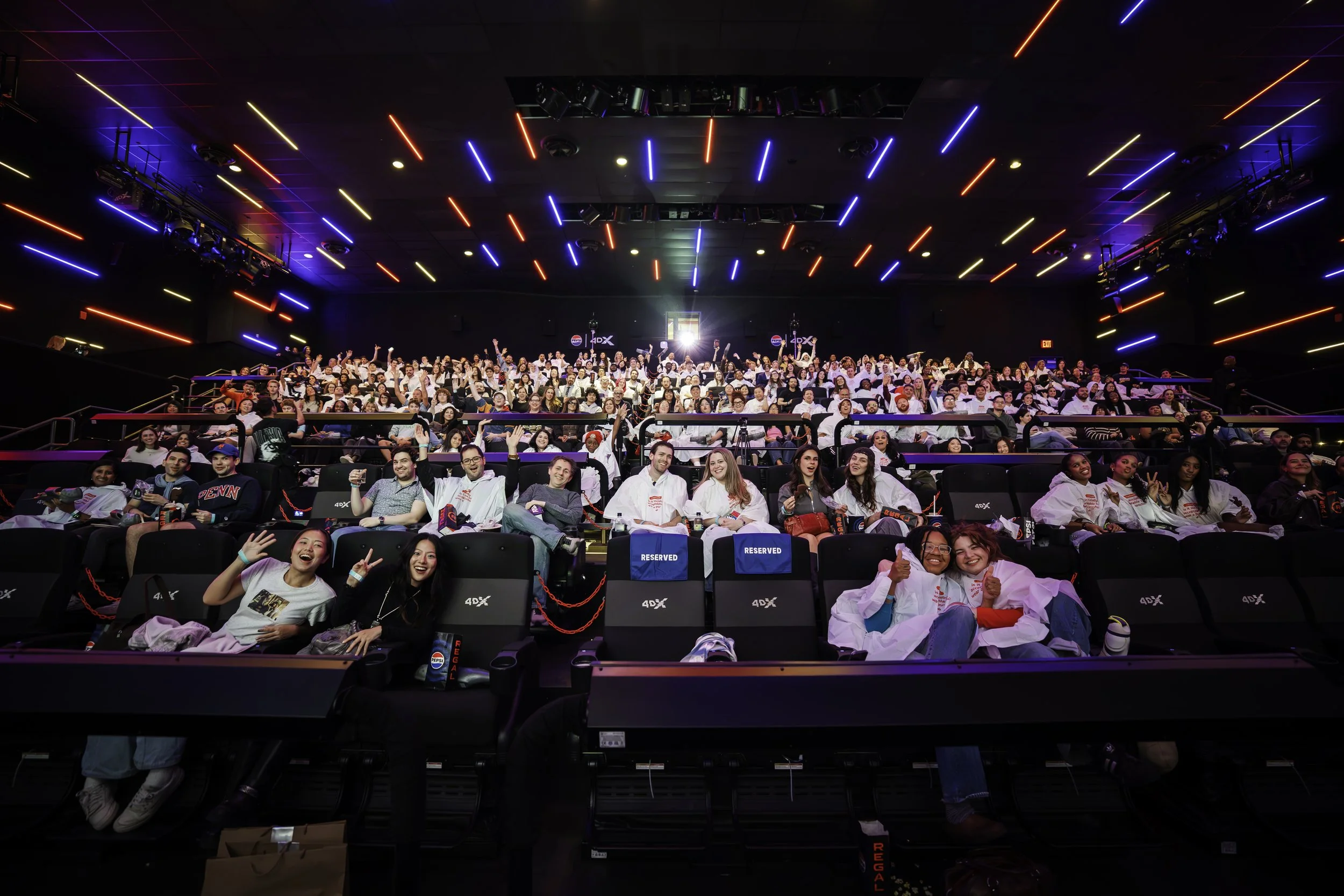 Packed cinema audience smiles and waves toward the camera beneath vivid red and blue ceiling lights, creating an energetic, celebratory mood.