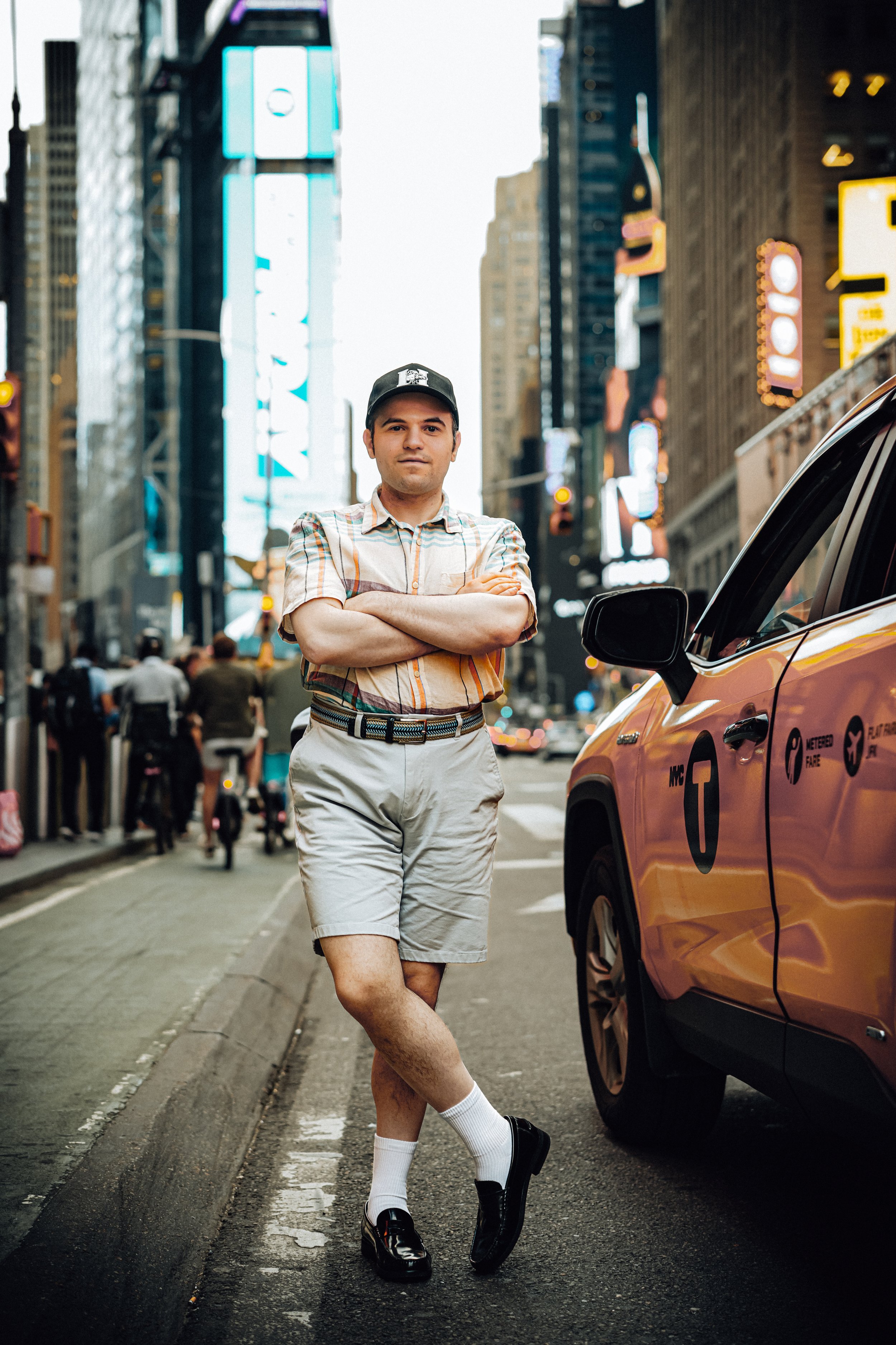A man in a plaid shirt, shorts, and a baseball cap stands confidently with arms crossed on a busy city street at dusk, with tall buildings, neon signs, and a yellow taxi cab nearby.