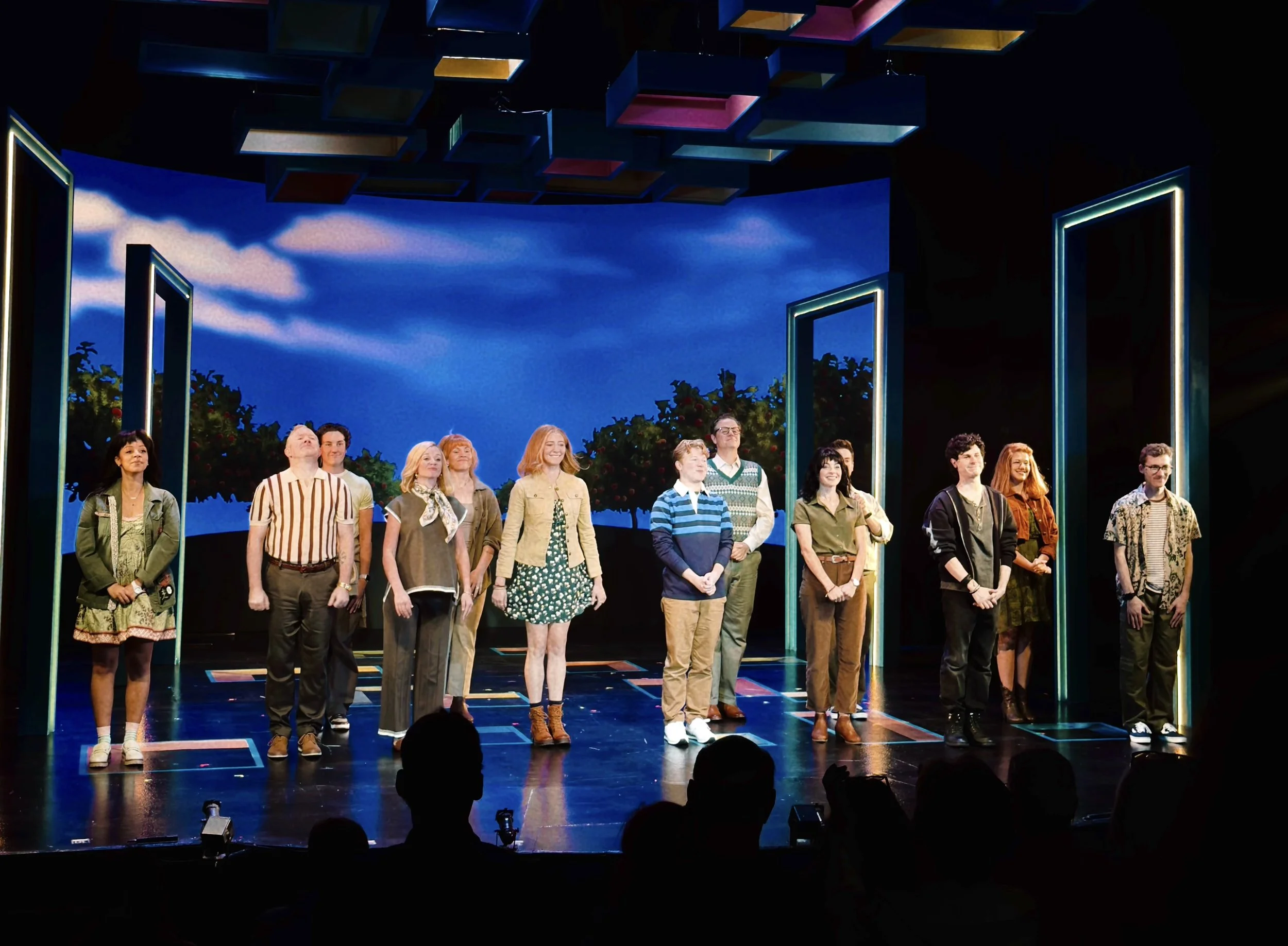 Diverse ensemble stands on a theater stage beneath colorful panels, framed by open doors, as audience silhouettes watch joyfully below.
