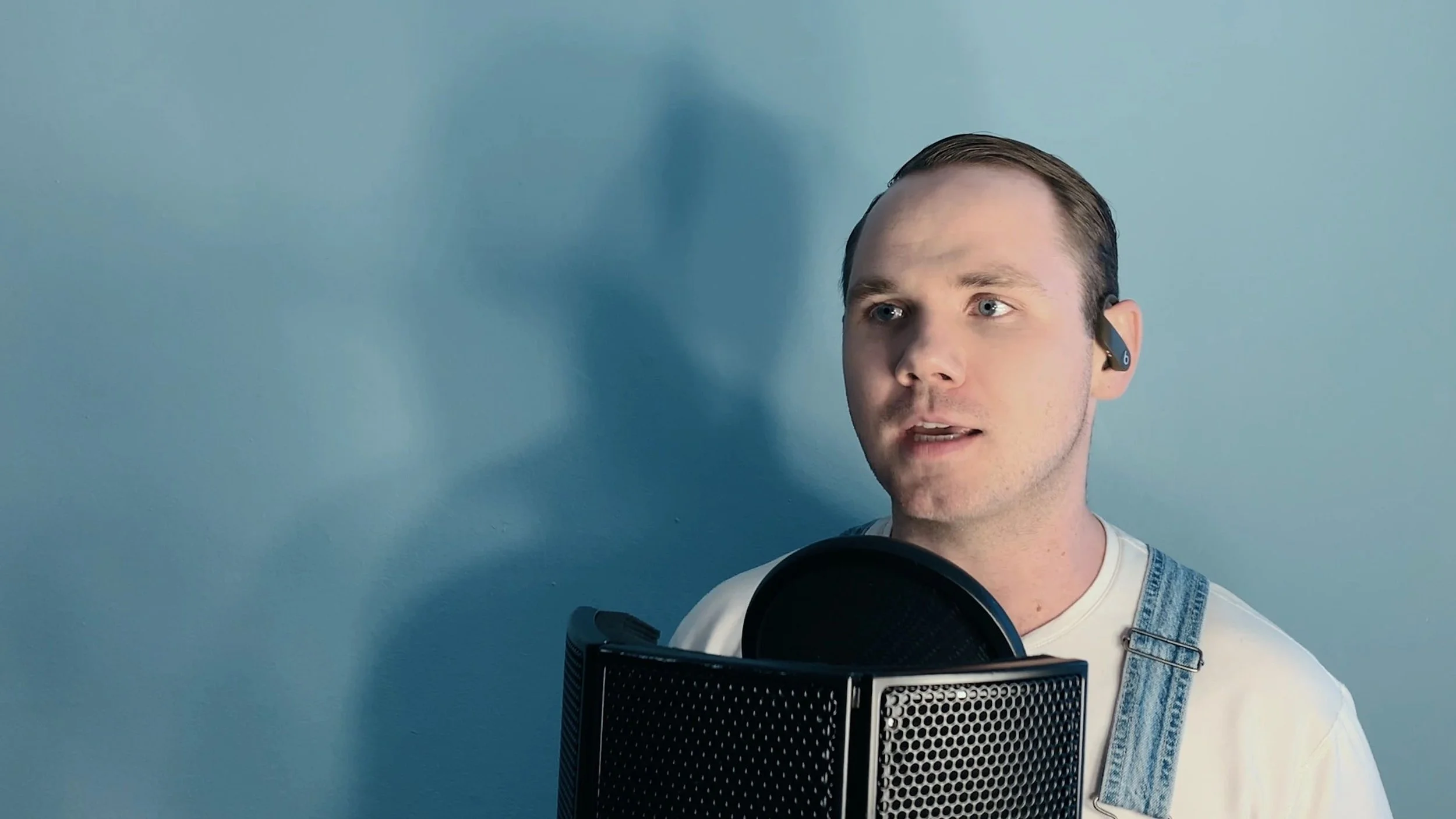 A man with short dark hair and light skin wearing overalls and a white t-shirt, singing into a microphone in a recording studio. He is also wearing a wireless earpiece.