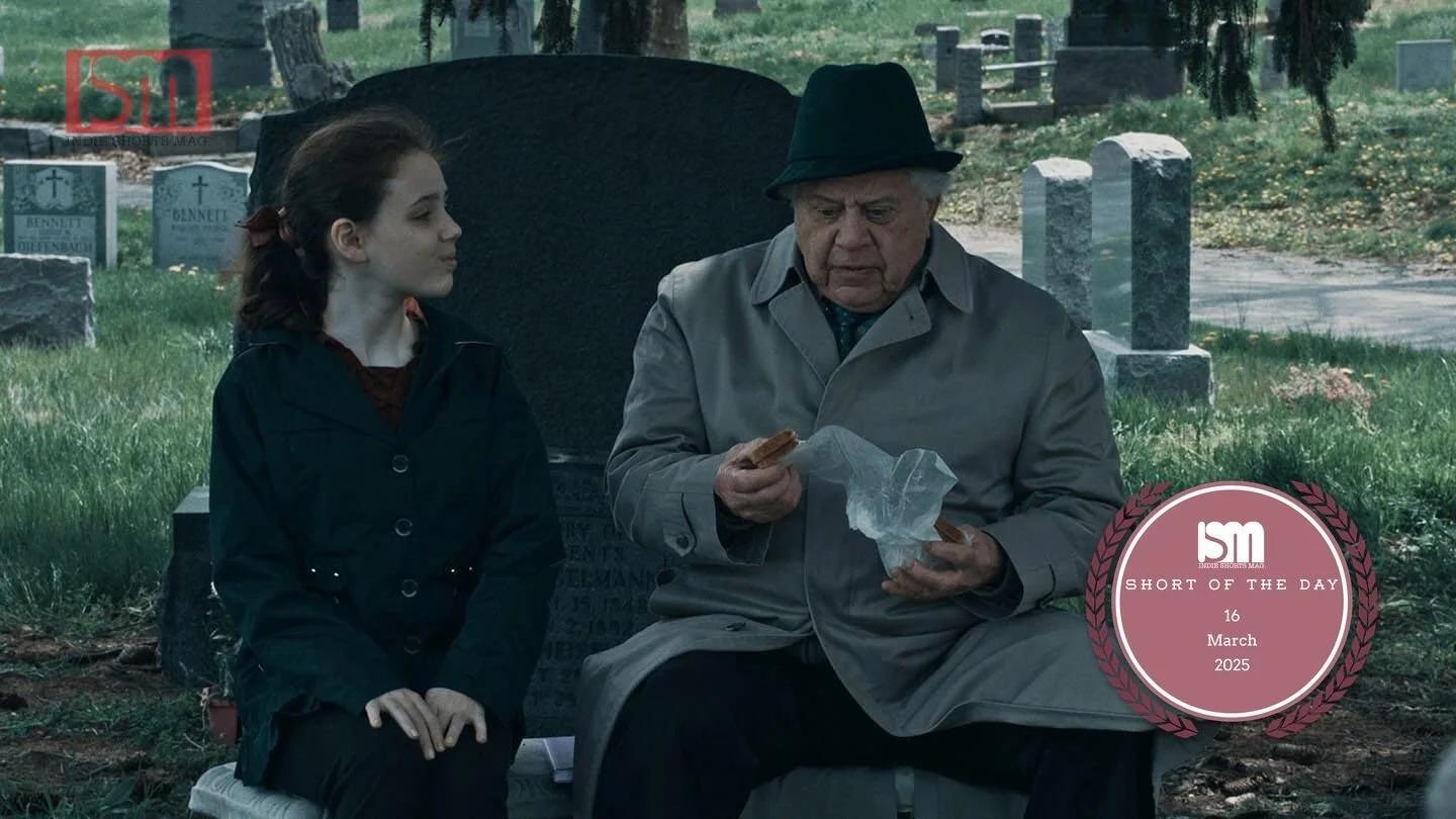 A young girl and an elderly man sitting on a bench in a cemetery, with headstones in the background. The man is wearing a gray trench coat and a black hat, eating a snack. The girl is wearing a dark jacket, looking at the man.