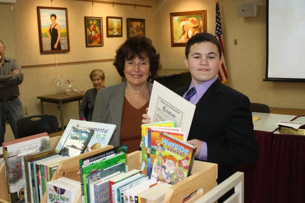 A woman and a young man are smiling and holding a certificate at a book event. There is a bookshelf filled with children's books in front of them. Background features framed paintings, an American flag, and several other people.