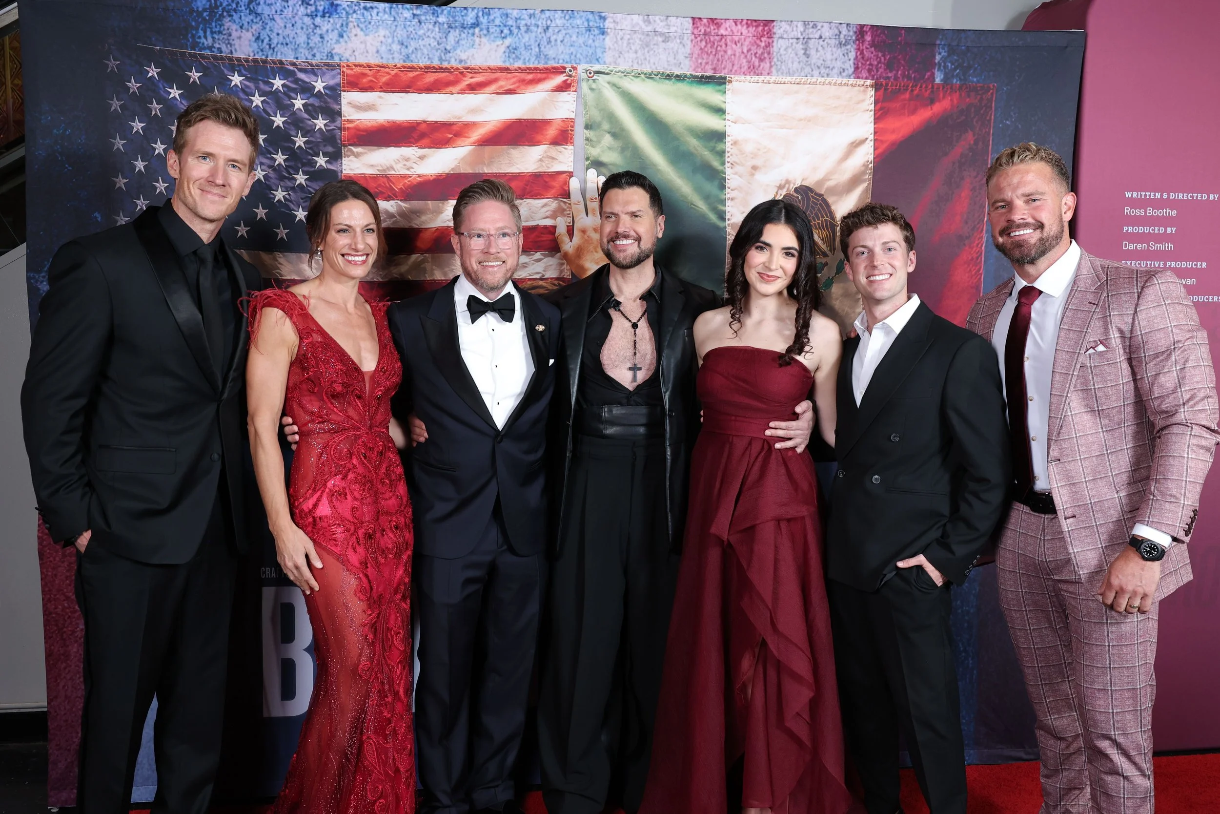 Seven adults pose in formal attire before a backdrop featuring U.S. and Mexican flags; the group smiles warmly at a festive event.