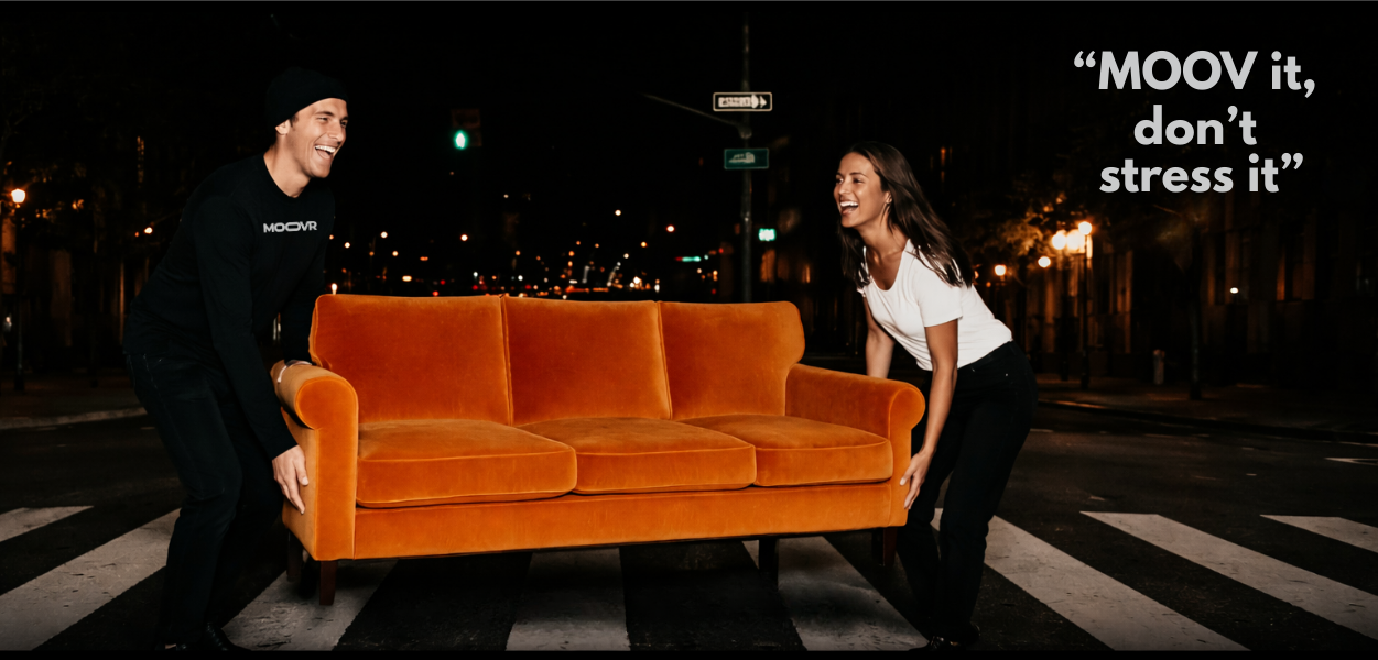 Two young adults, a man and a woman, are smiling and lifting a vintage orange sofa on a city street at night. The scene is illuminated by streetlights, and there is a signpost and traffic lights in the background. The image includes the quote 'MOOV it, don't stress it' in the top right corner.