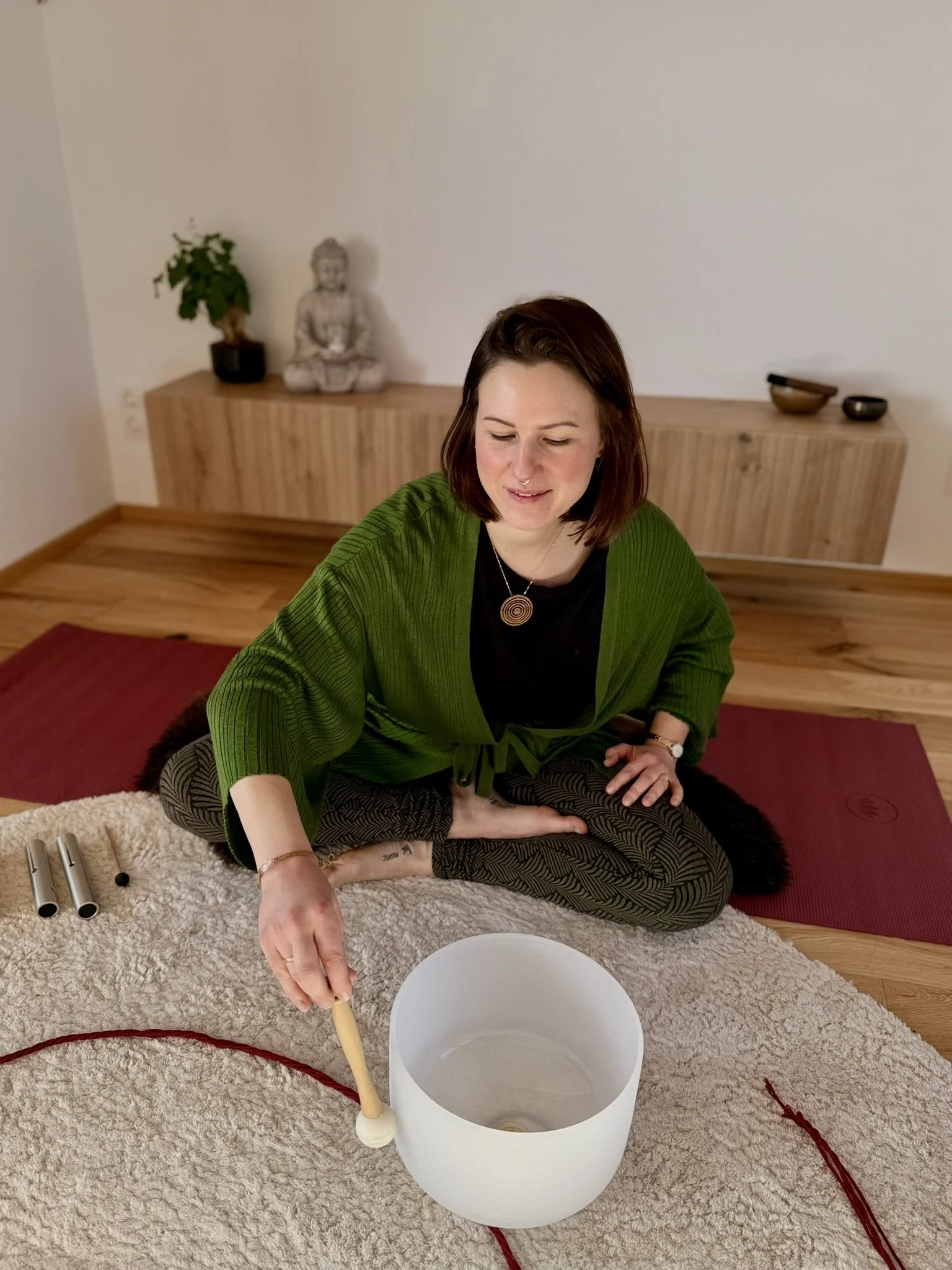 Eine Frau sitzt im Schneidersitz auf einem Teppich in einem Raum, hält einen Schlägel und spielt eine Tibetanische Klangschale.