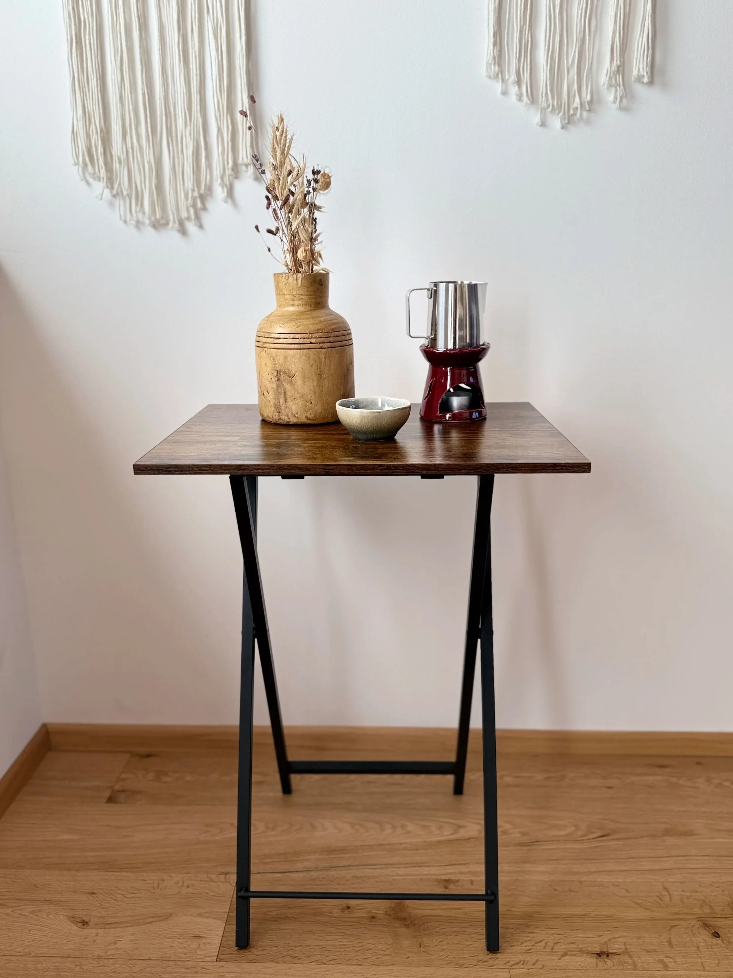 Ein Holztisch mit schwarzen Metallbeinen, auf dem eine Vase mit Trockenblumen, eine kleine Schale und eine Kaffeemaschine stehen, vor einer weißen Wand mit Wandbehängen aus Makramee.
