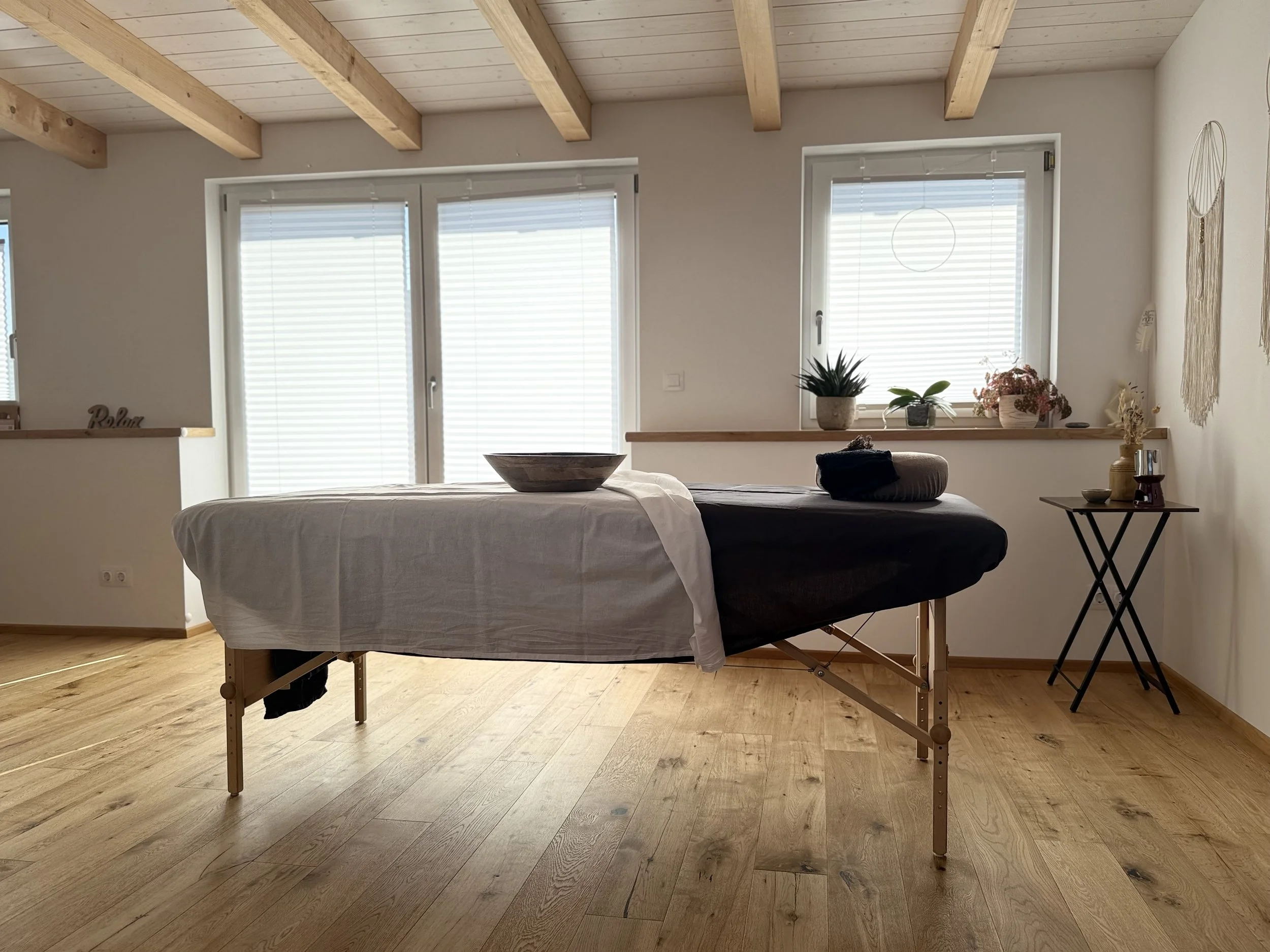 Ein Massagetisch in einem hellen Raum mit Holzbalkendecke, Fenster mit Rollläden, Dekoration an der Wand und Pflanzen auf der Fensterbank.