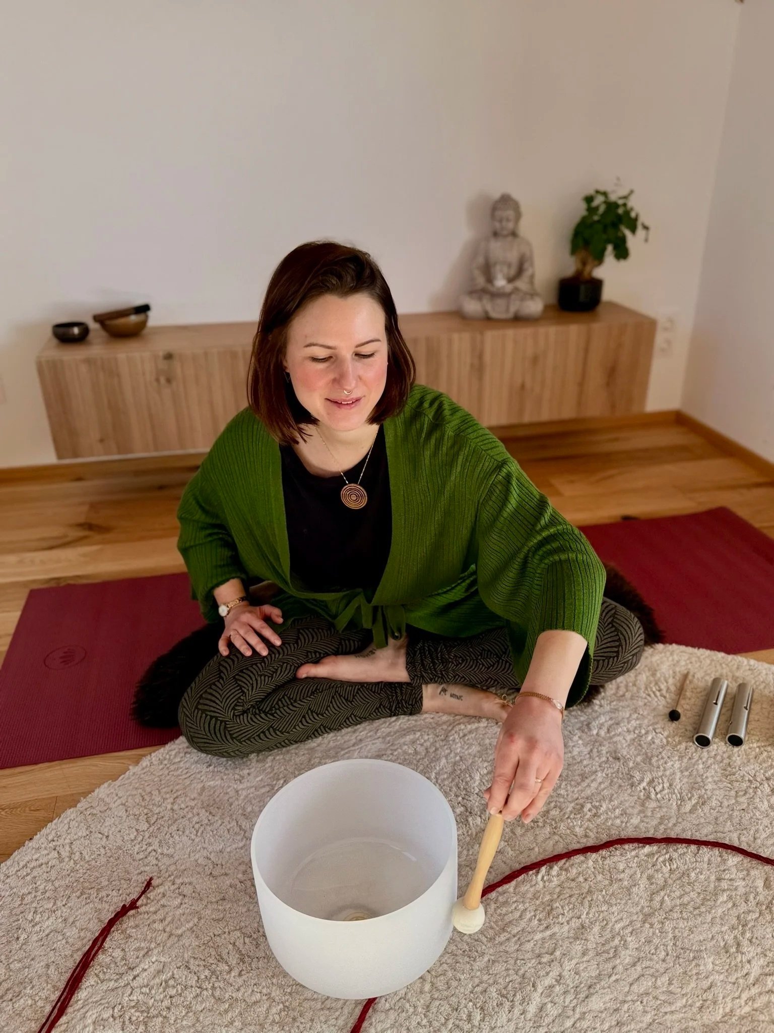 Eine Frau sitzt im Schneidersitz auf einem Teppich und spielt eine Hinduspiel mit zwei Trommeln. Vor ihr steht eine große Keramikschüssel, und um sie herum liegen rote Wäsche zu einem Kreis gerollt.