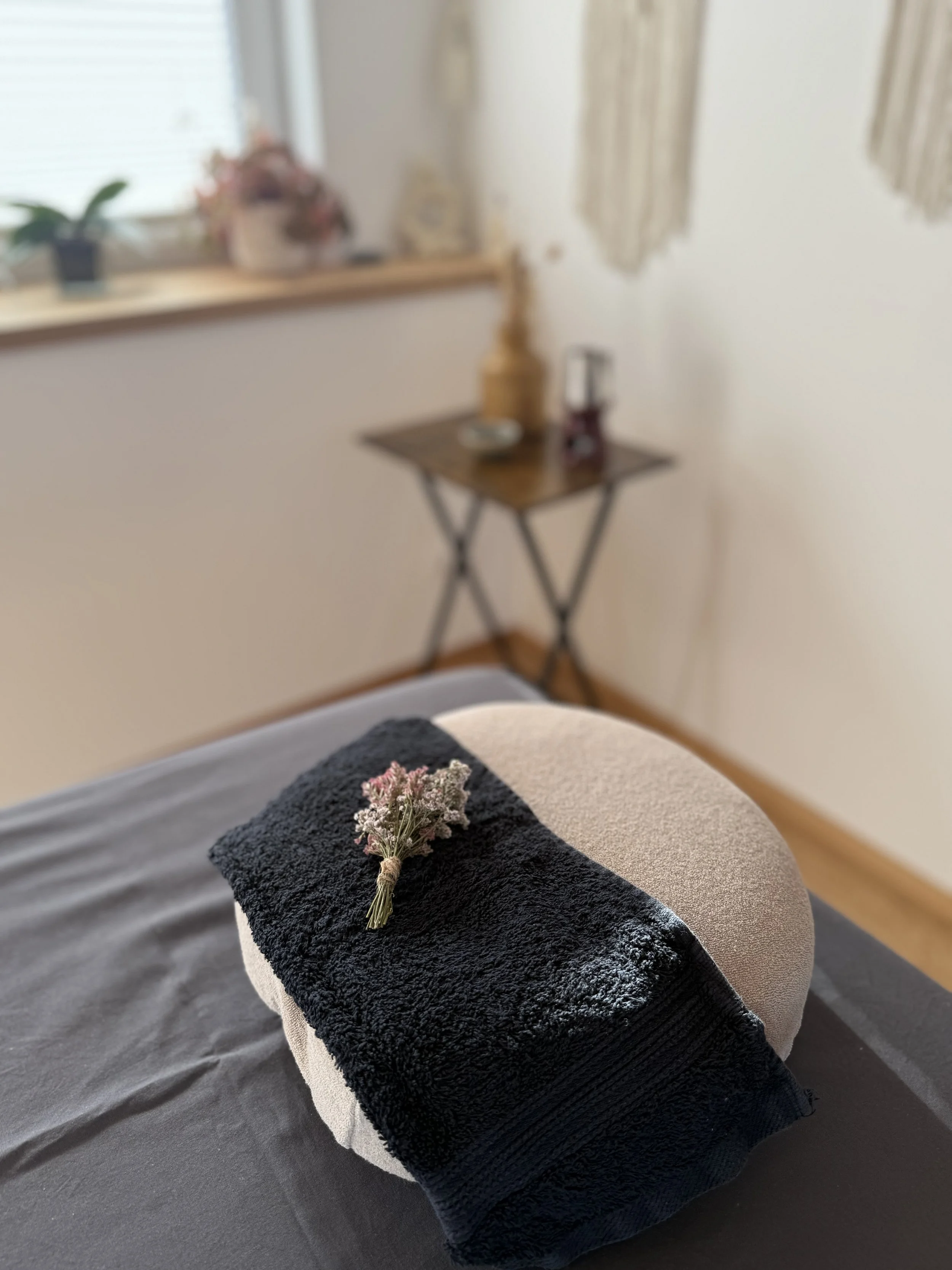 Im Vordergrund und in Nahaufnahme ist das Kopfteil einer Massageliege zusehen. Darauf ein Massagekissen, dekoriert mit rosa und lila Trockenblumen. Im Hintergrund ist ein kleiner Tisch zu sehen, auf dem ein Meersalz-Schüsselchen, Teelicht, Massageöl