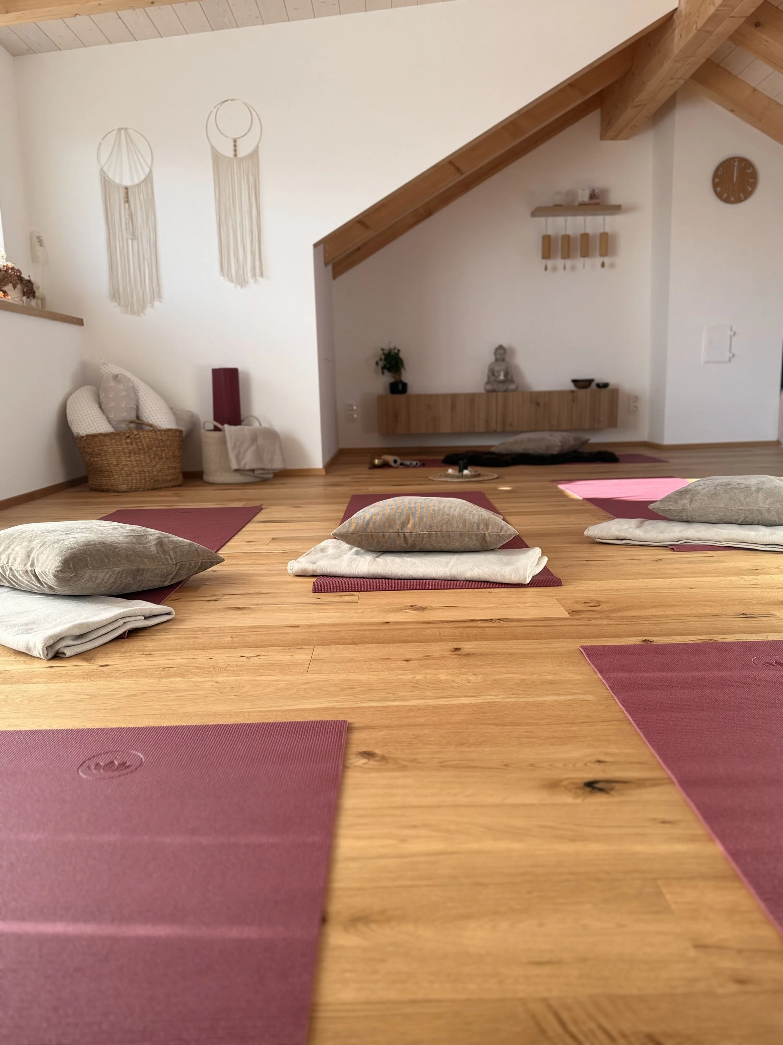 Yoga-Raum mit gestapelten Yogamatten, Kissen und Decken auf Holzboden, Wand mit Dekorationen, wie Traumfänger, und einer kleinen Buddha-Statue, schräges Holzdecke.