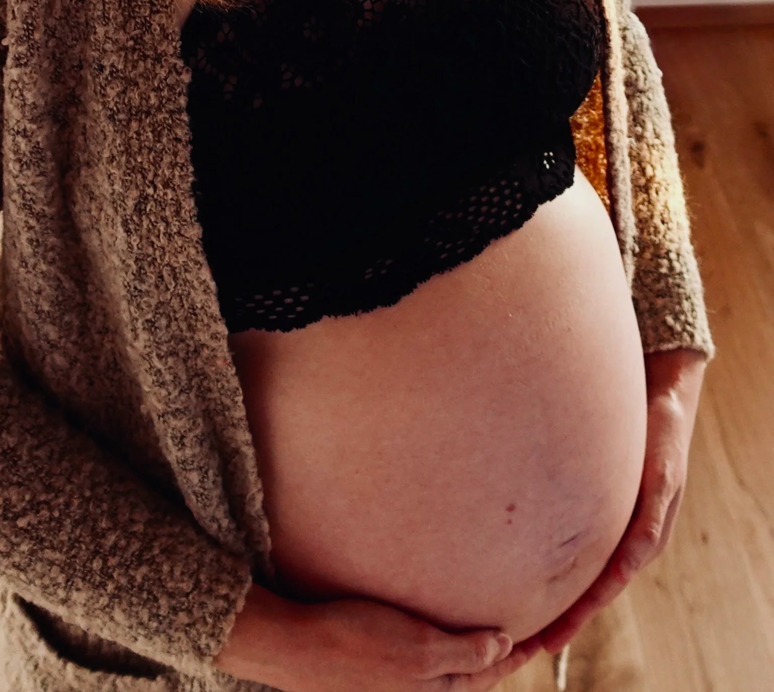 Nahaufnahme von einer schwangeren Frau. Im Fokus ist der freie Babybauch, sie hält ihre Hände liebevoll am unteren Bauch. Sie trägt ein schwarzes Bustier und eine beige offene Wolljacke.