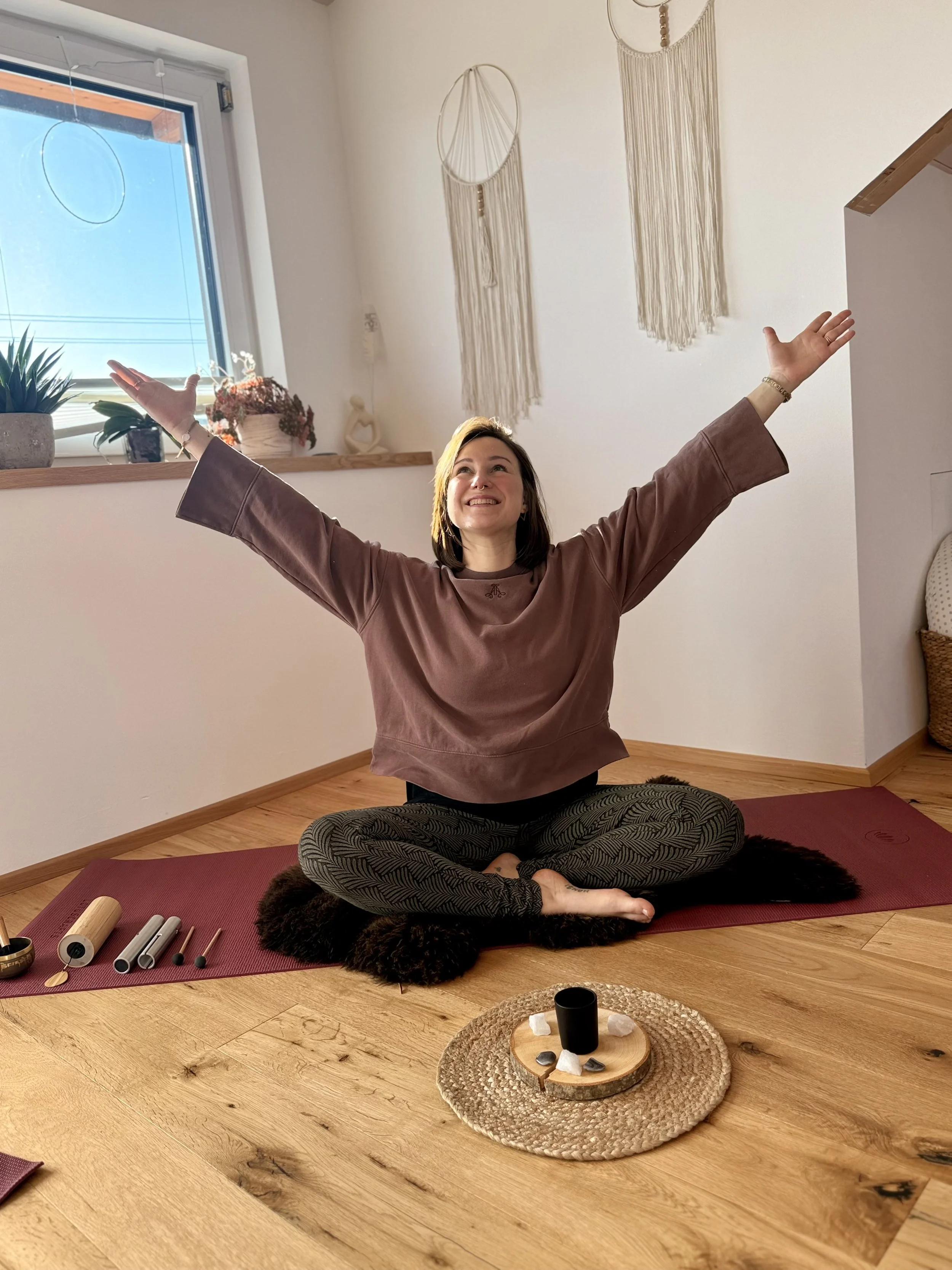 Doula Dé sitzt auf Yogamatte mit Klanginstrumenten daneben und streckt Hände in 60 Grad Winkel nach oben und lächelt