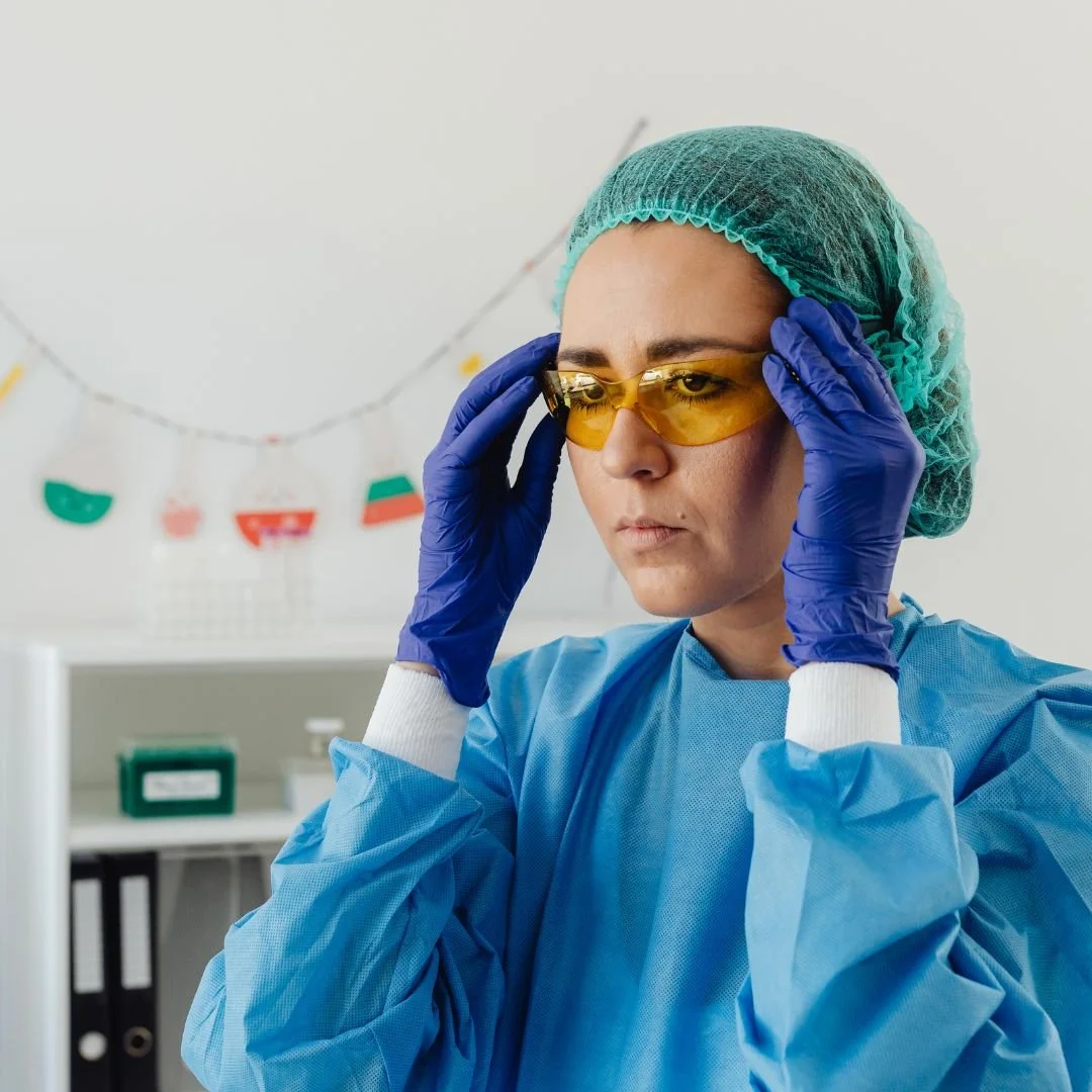 A female healthcare worker in medical scrubs, wearing a surgical cap, yellow safety glasses, and blue gloves, adjusting her glasses in a medical setting with colorful decorations in the background.