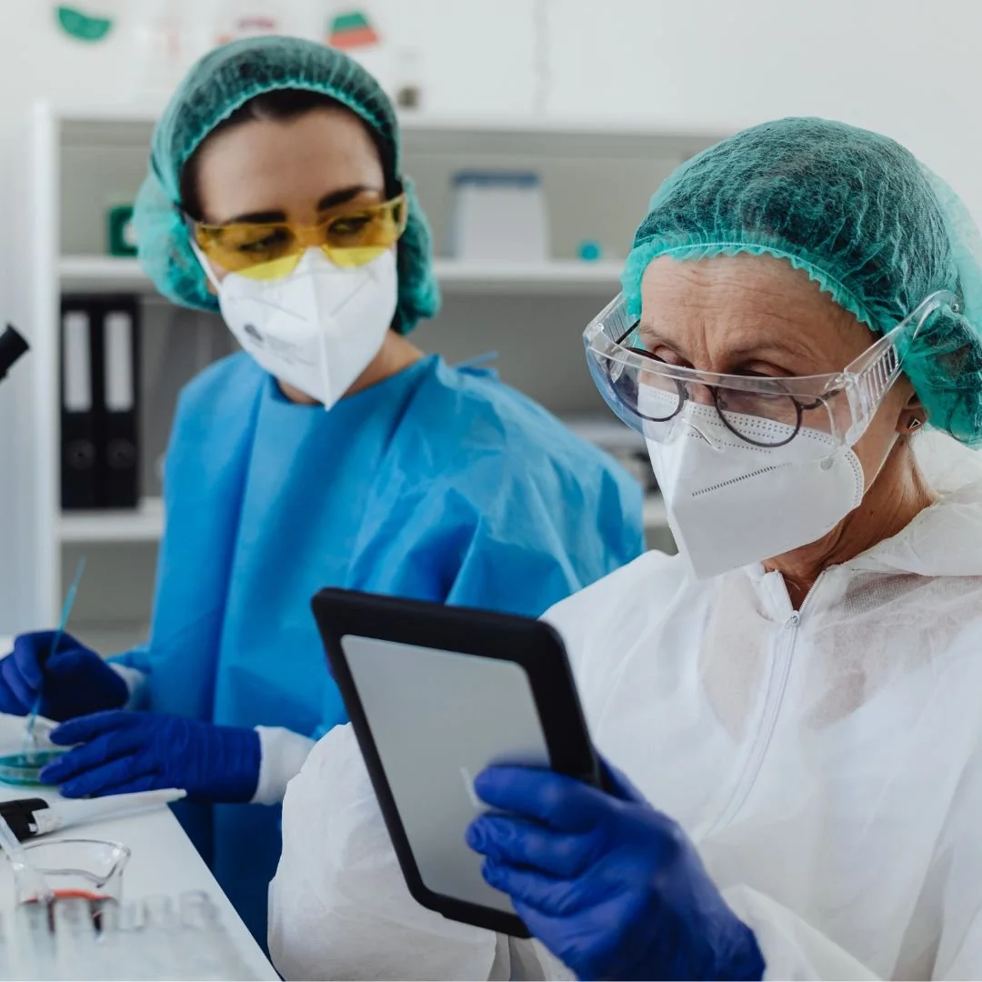Two medical professionals wearing protective gear, including masks, goggles, gloves, and scrubs, working in a laboratory or medical setting.
