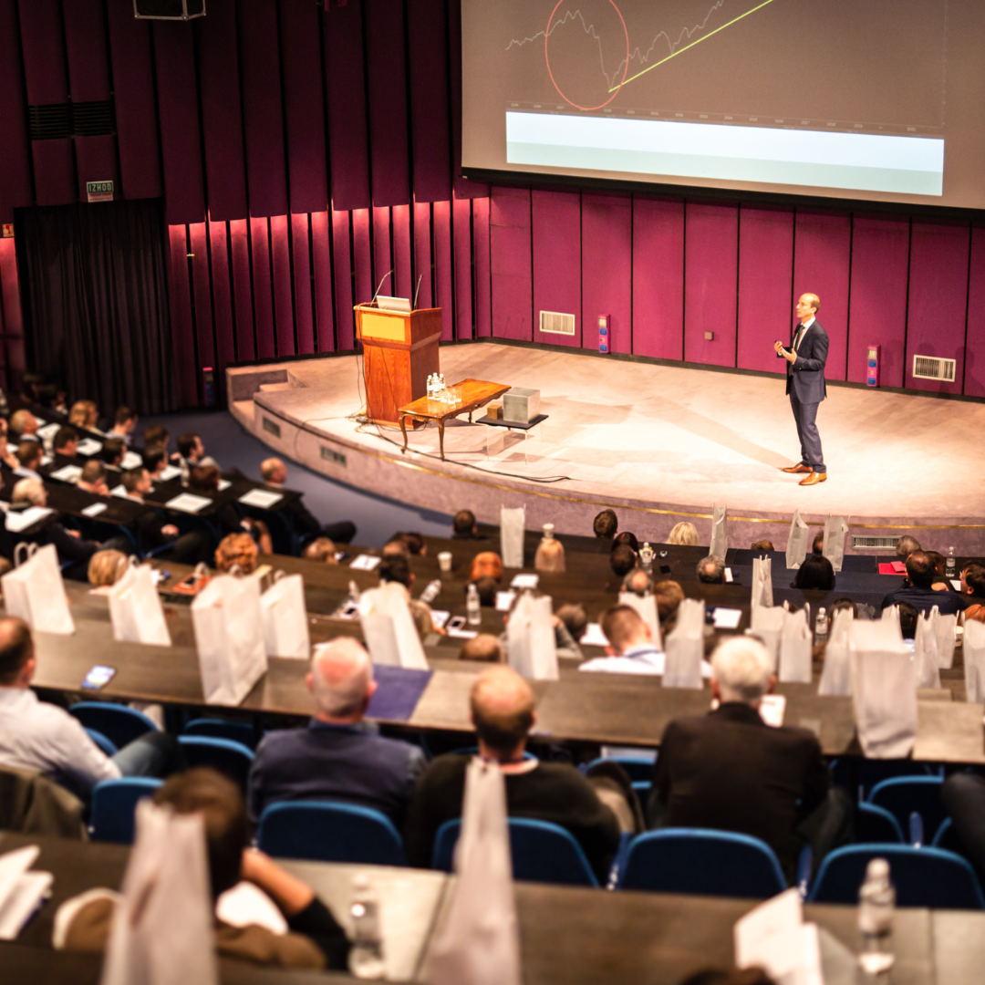 A man in a dark suit giving a presentation in an auditorium with purple walls, an audience seated at tables with water bottles, and a large screen displaying a graph with financial lines.