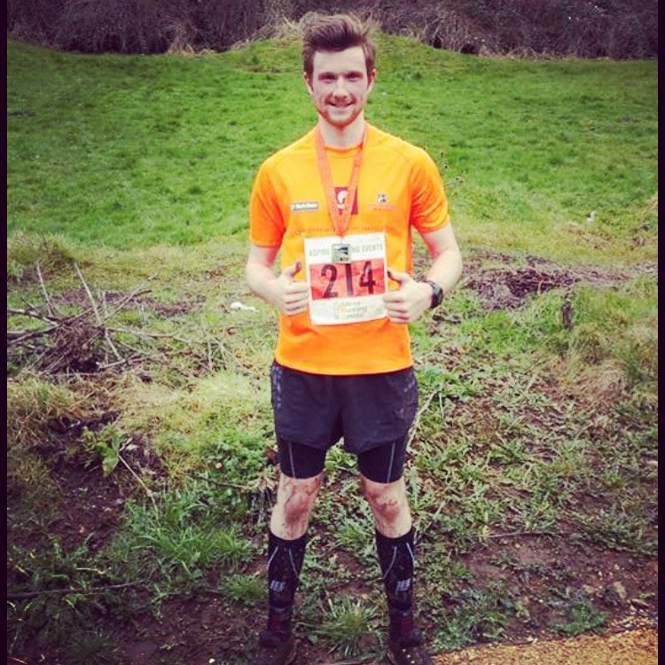 A young man in an orange athletic shirt and black shorts, wearing a race bib with the number 24 and a medal around his neck, standing outdoors on a grassy trail with muddy patches and green hills in the background.