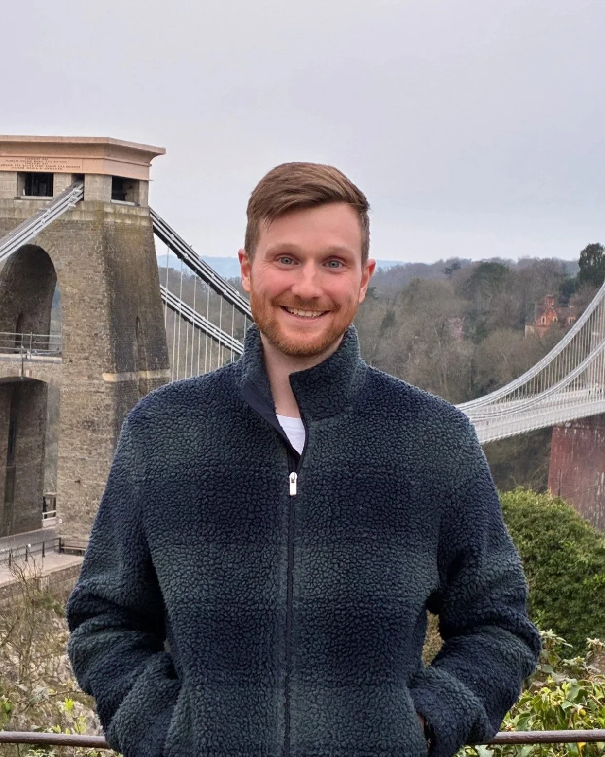 A man smiling outdoors in front of the Clifton Suspension Bridge in Bristol, England, wearing a navy fleece jacket with hills and trees in the background.