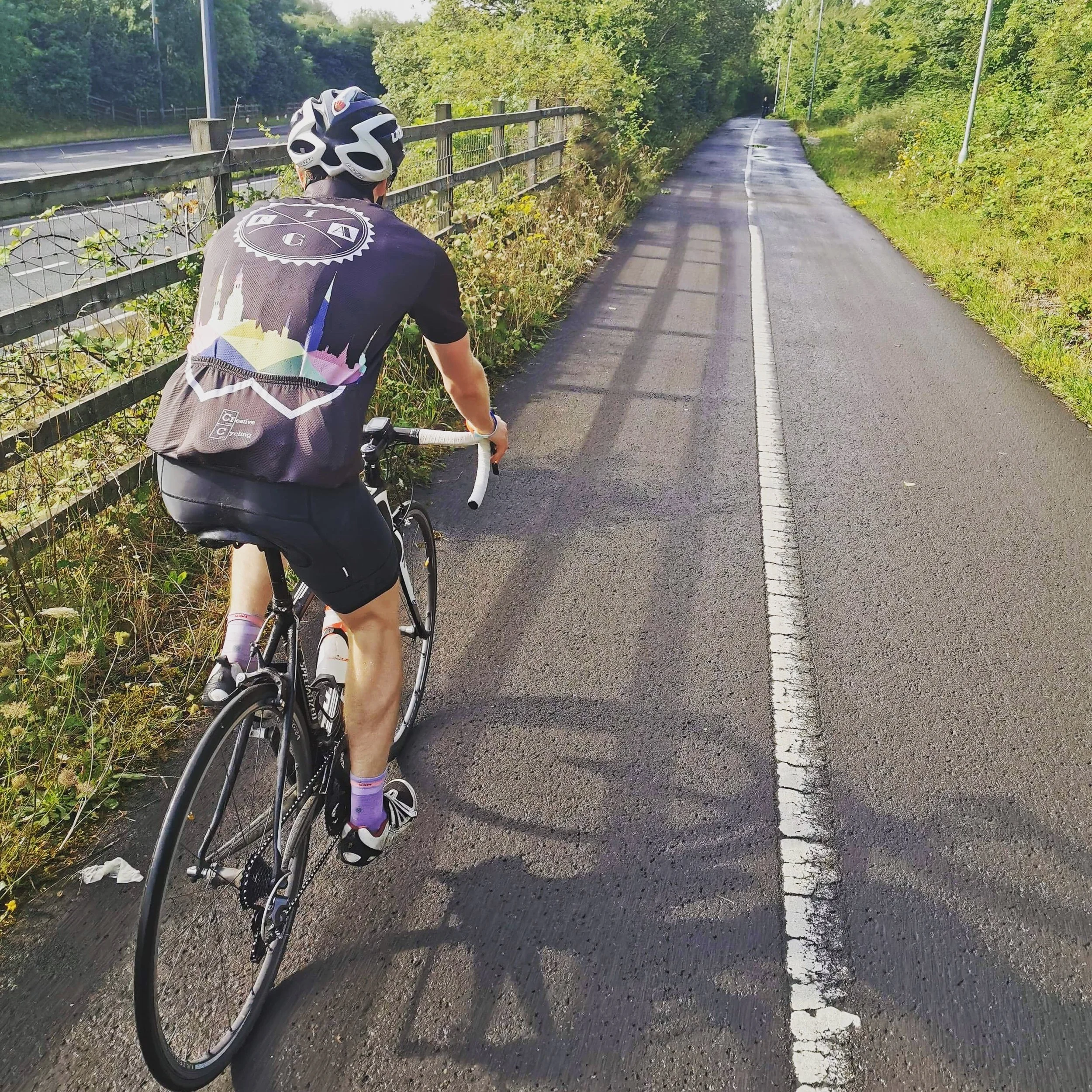 A cyclist wearing a helmet and cycling gear rides along a paved bike path bordered by greenery and a fence, with a road visible in the background.