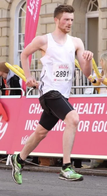 A man running in a race, wearing a white tank top, black shorts, green running shoes, and a race bib number 2891, on a city street with spectators and race banners in the background.