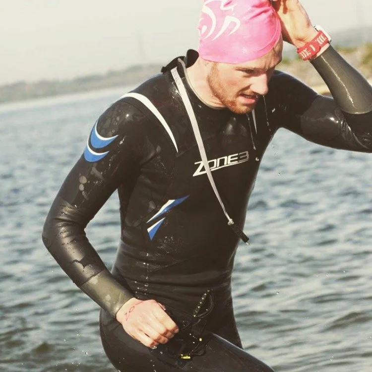 A man wearing a pink swim cap and a black wetsuit with blue and white accents, standing in water near a shoreline.